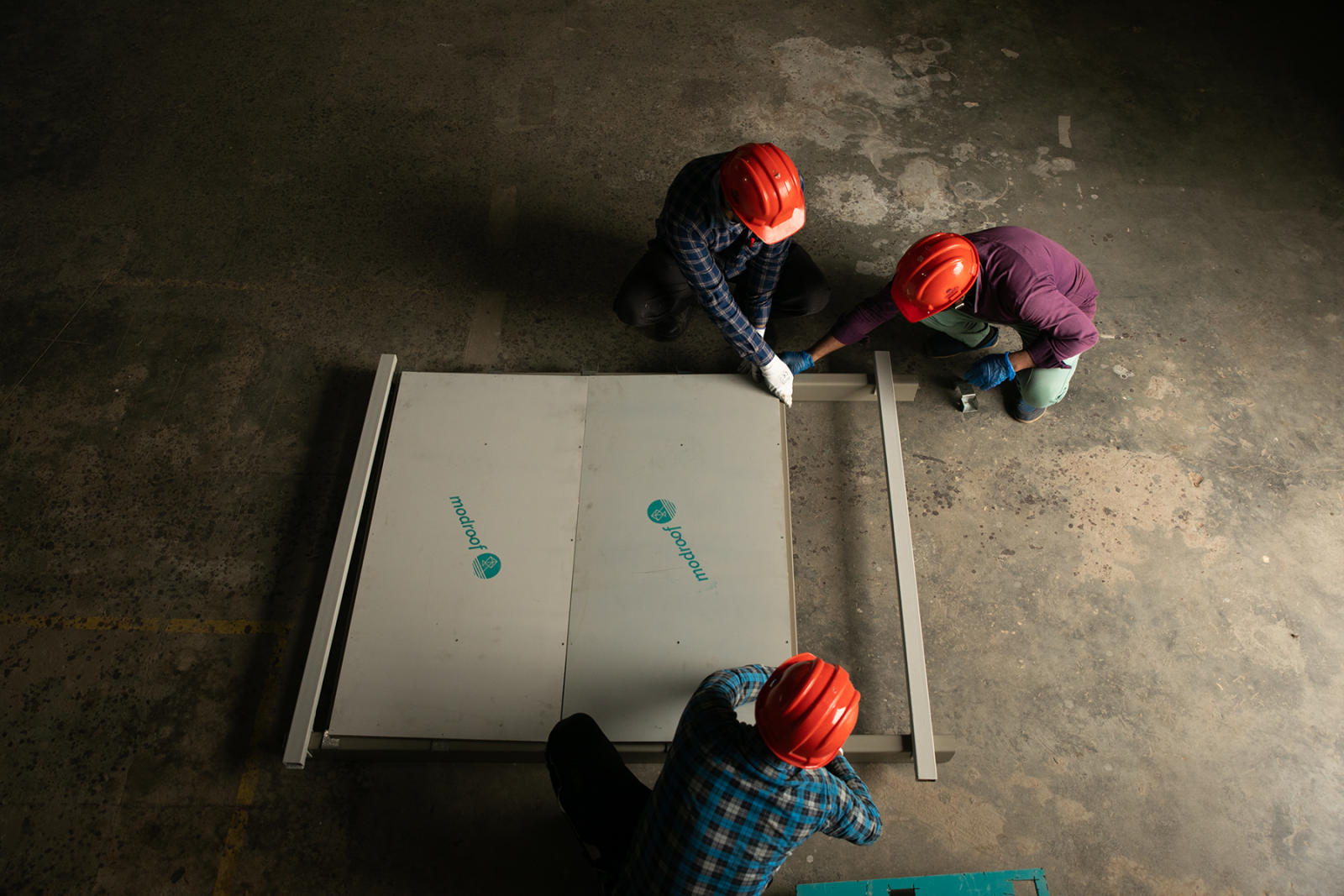 This is a demonstration of ModRoof panels being assembled. This is an aerial shot of the panels on a concrete floor with three people in orange hard hats sitting and working.
