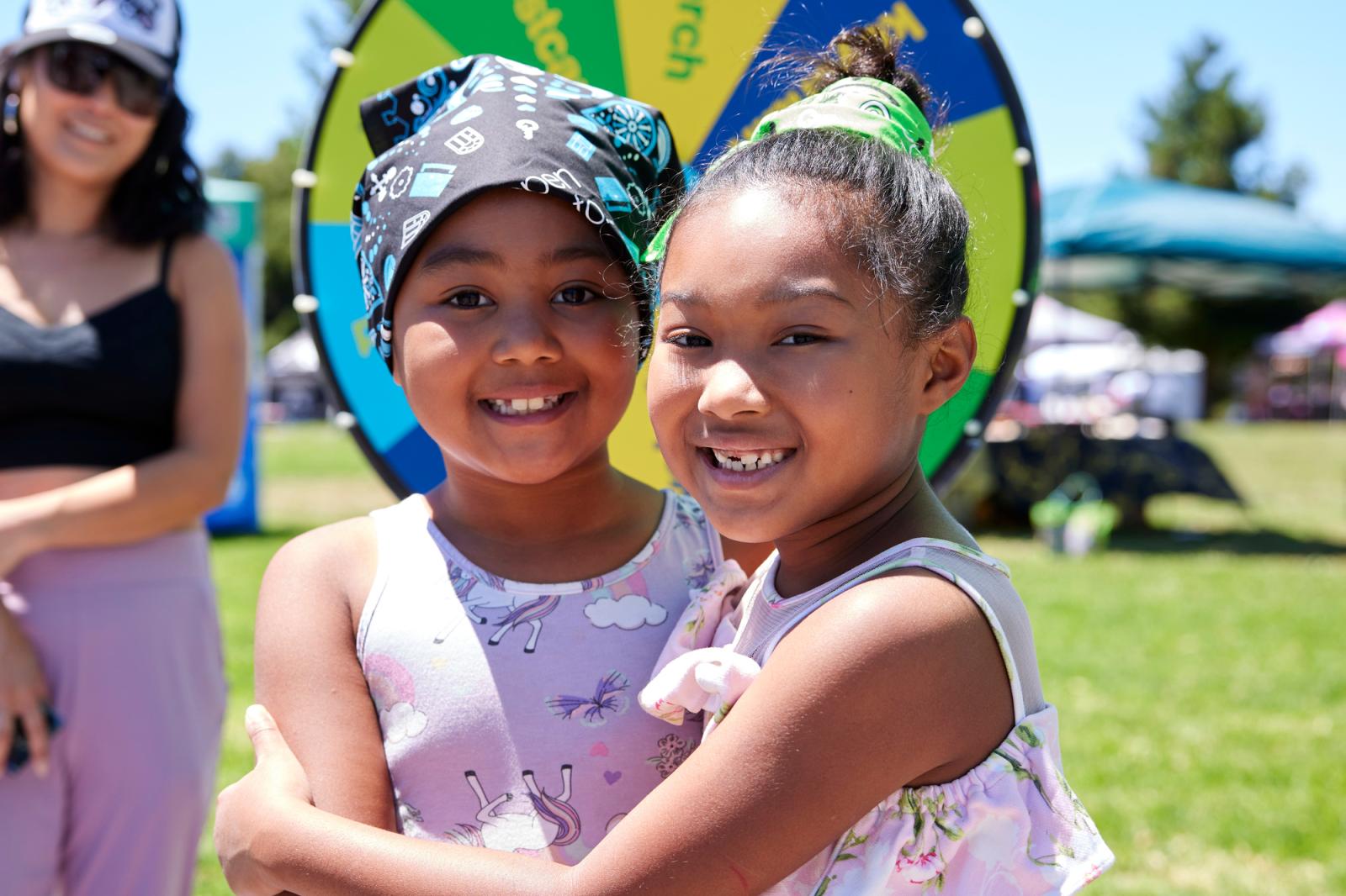 A mom smiling as two young girls in pink shirts pose for a photo in front of a prize wheel.