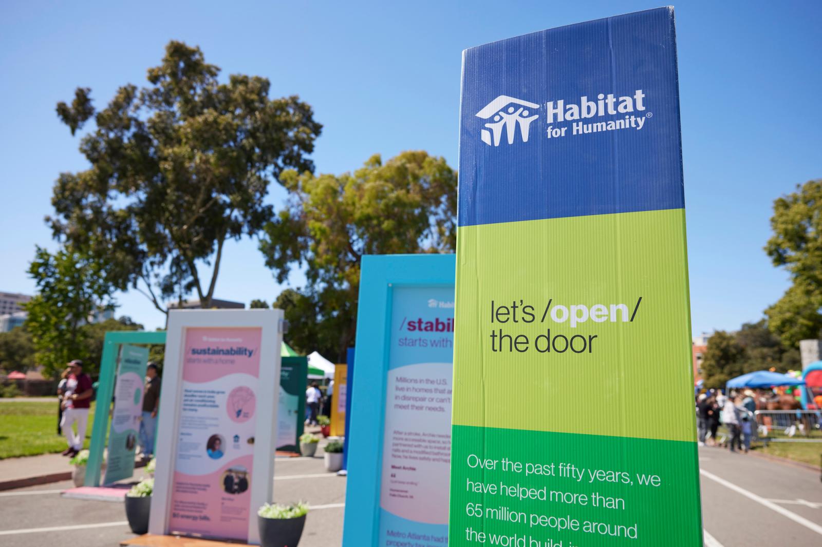 Exhibit doors in the background and a sign in the foreground that has the Habitat logo and says Let's Open the Door.
