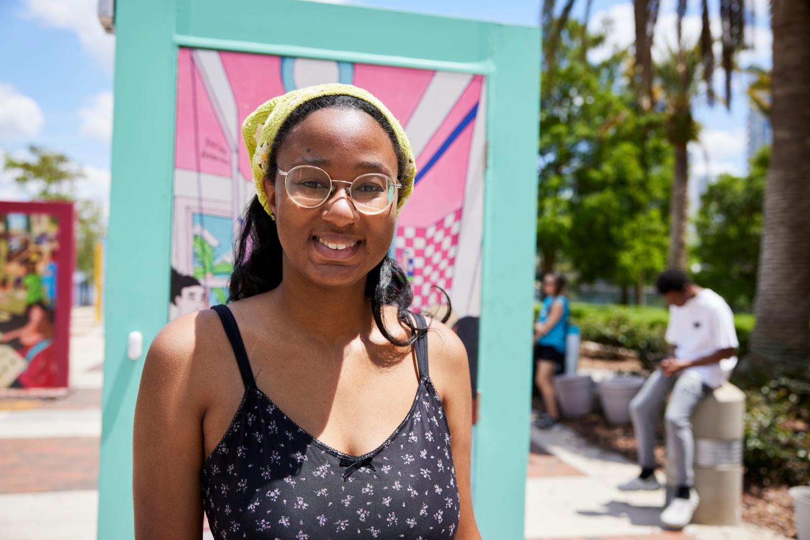 Woman in a black dress with white flowers, glasses and a yellow bandana in her hair posing in front of a door with a mint green door frame.
