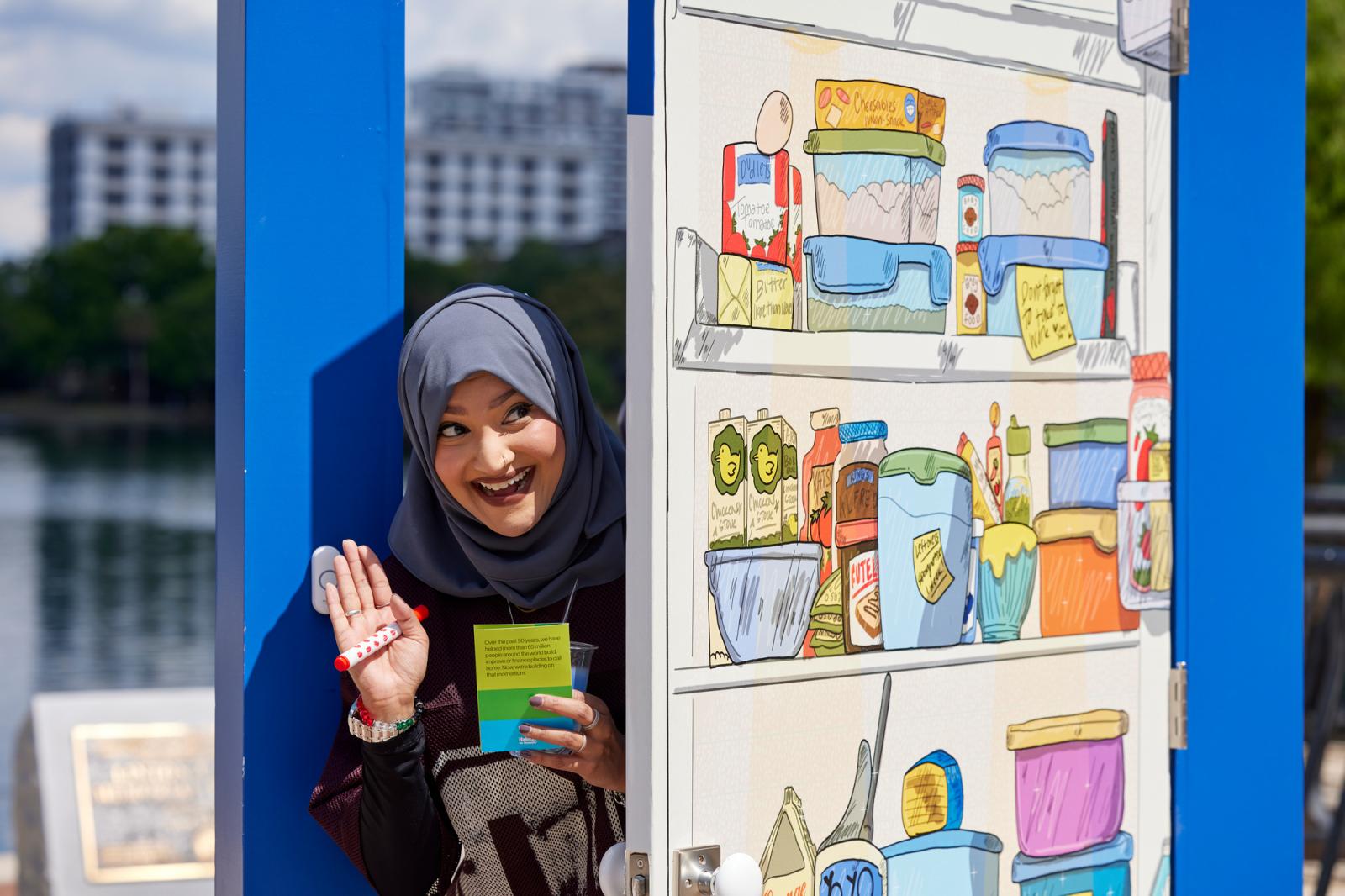 Woman in a gray hijab posing peeking through a dark blue door with an illustration of a fridge on it.