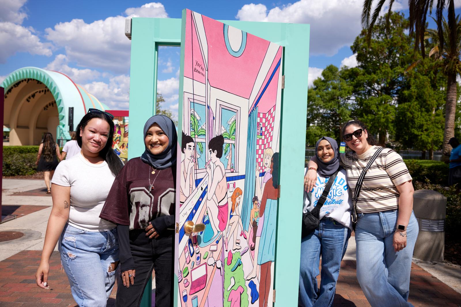 A group of four women, two with hijabs, posing in front of the illustrated door by the Orlando artist, Danniell Fennell.