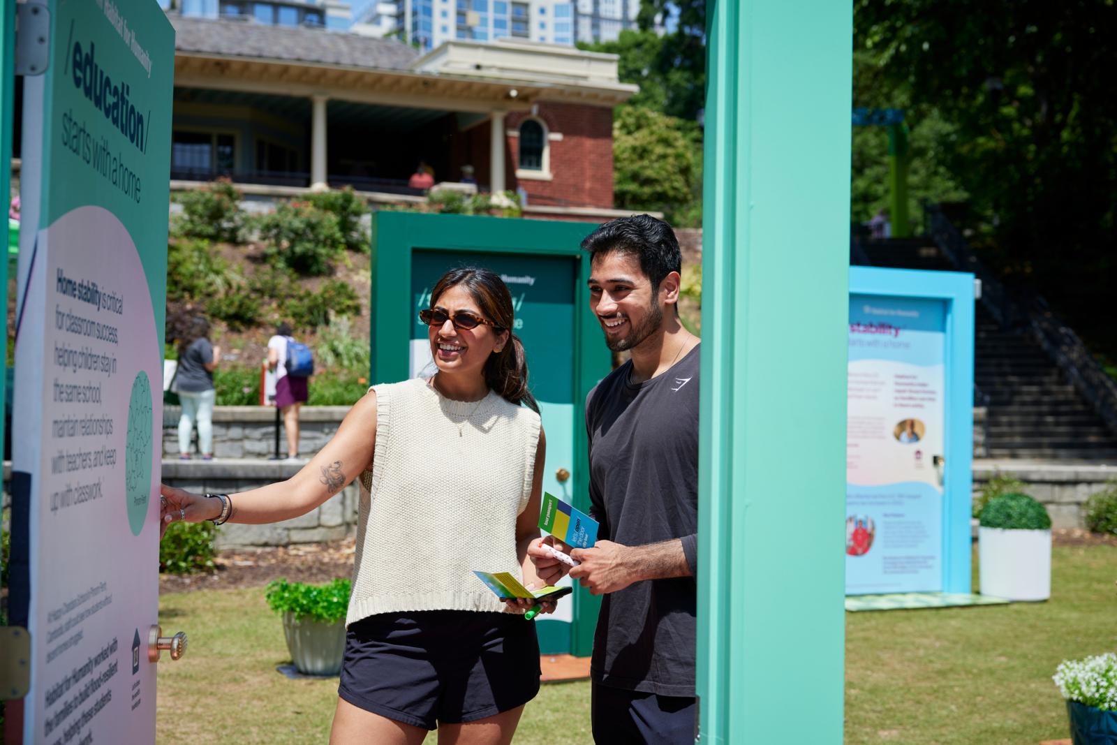 A woman in a white tank and black shorts next to a man in all black viewing a teal door.