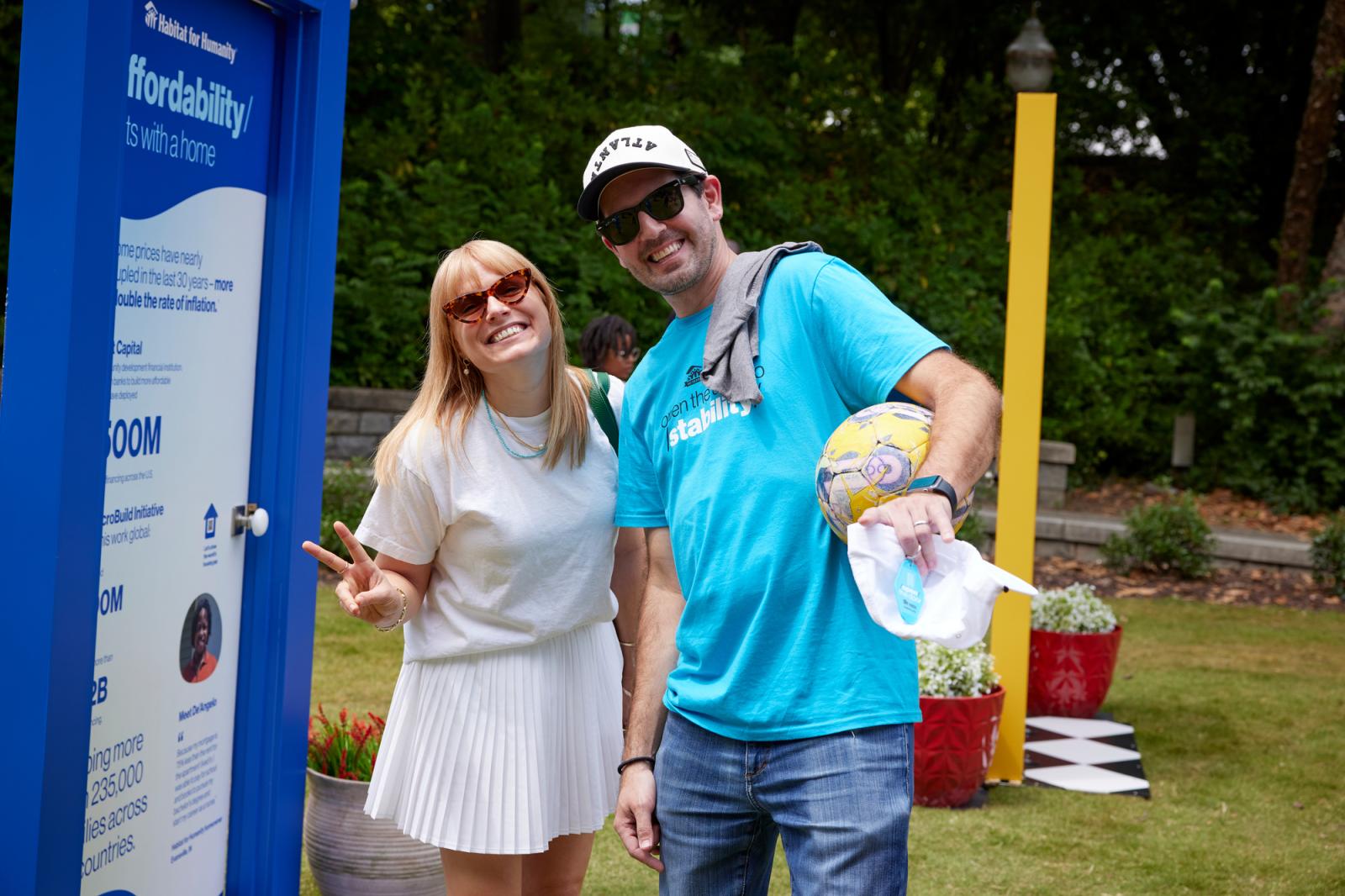 Woman in a white shirt and skirt posing with a man in a blue habitat shirt and ball cap next to a door with a blue frame.