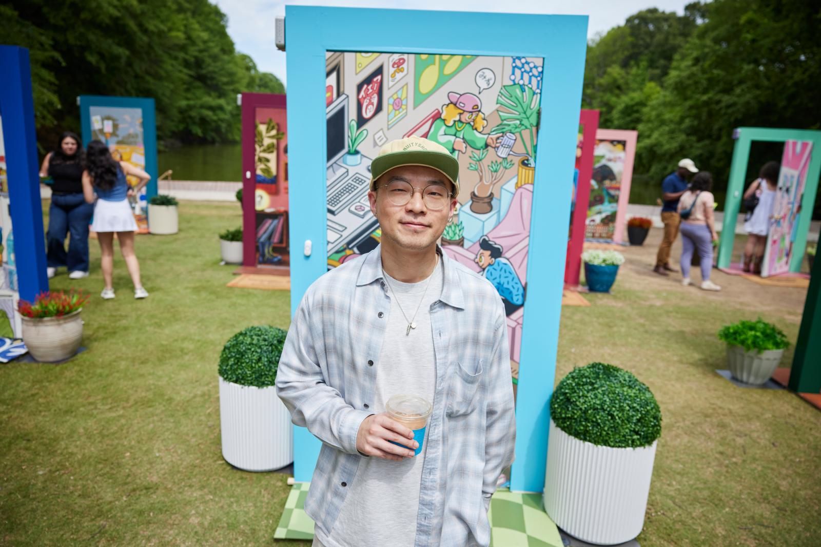 Man in checkered blue and white shirt with a white undershirt, glasses and a hat standing in front of a blue door with an illustration.