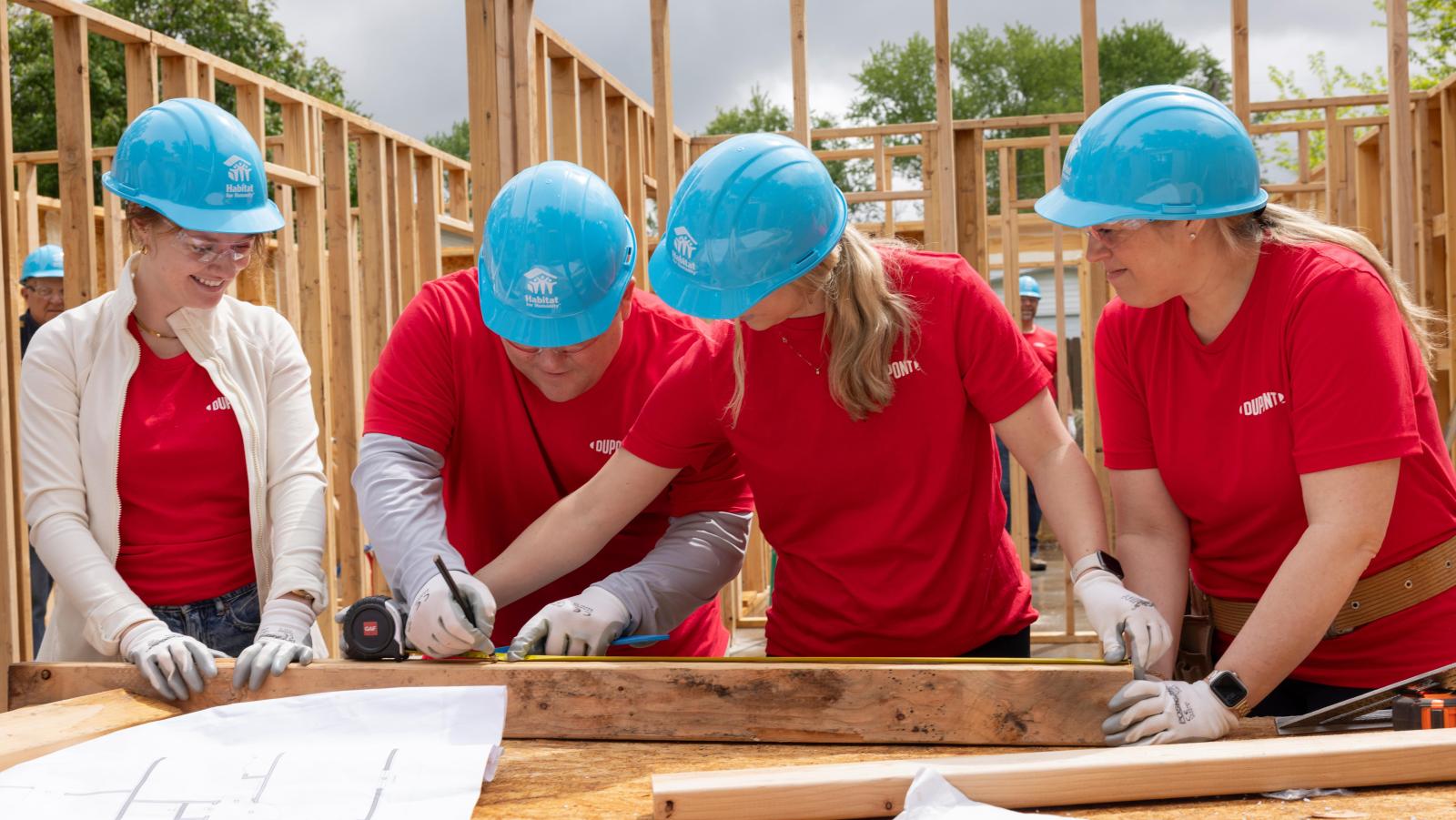 Four DuPont volunteers in red t-shirts and blue Habitat hard hats work on the site of a Habitat house.