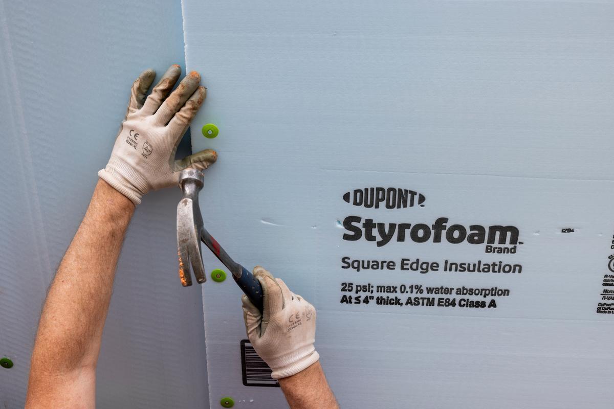 A DuPont volunteer's gloved hands hammering into a sheet of Styrofoam insulation.
