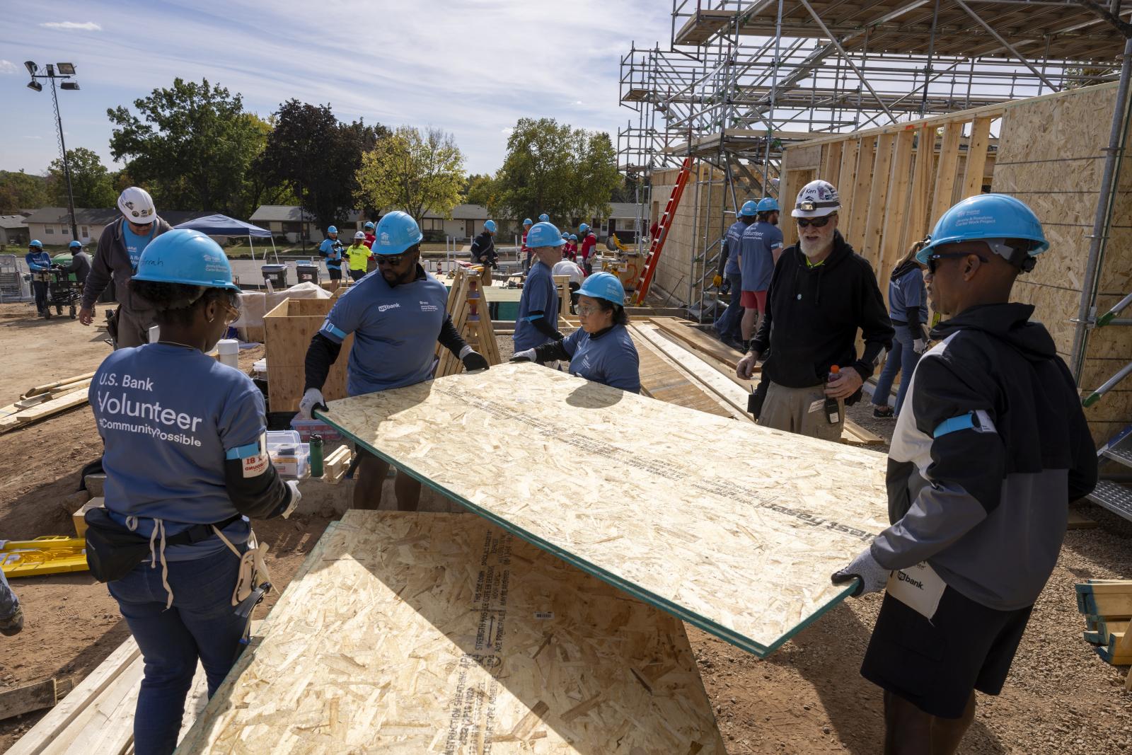 US Bank employees in bright blue hard hats, blue shirts and jeans carrying a piece of wood on a job site.