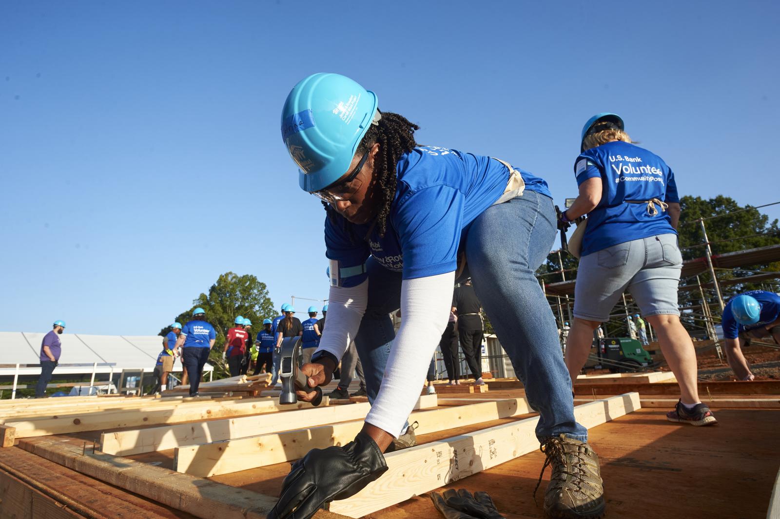 US Bank employee lifting wood on a job site in a bright blue hard hat, blue shirt and jeans.