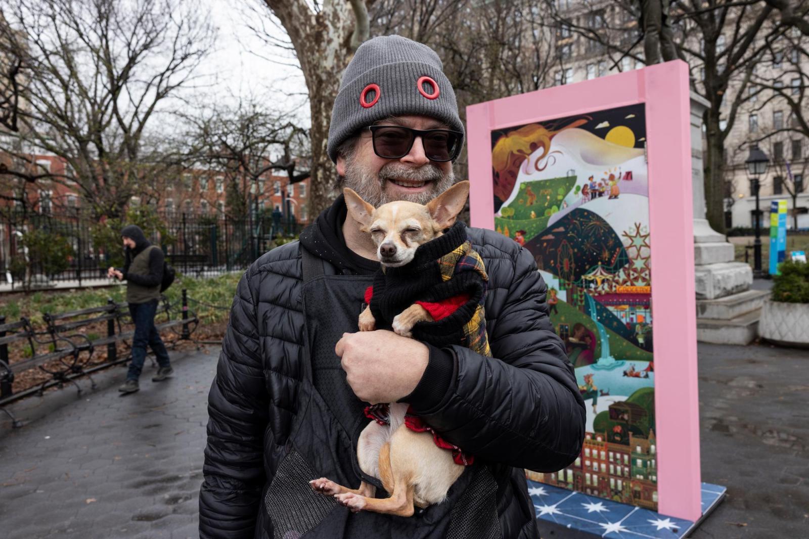A man with a beanie, sunglasses and a black rain jacket holding a dog in a coat in front of a door.