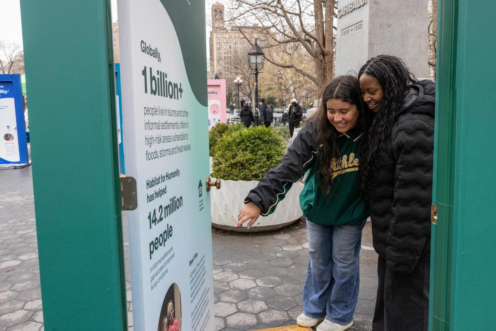 Two women with black hair and black raincoats viewing a teal door with stats about housing.
