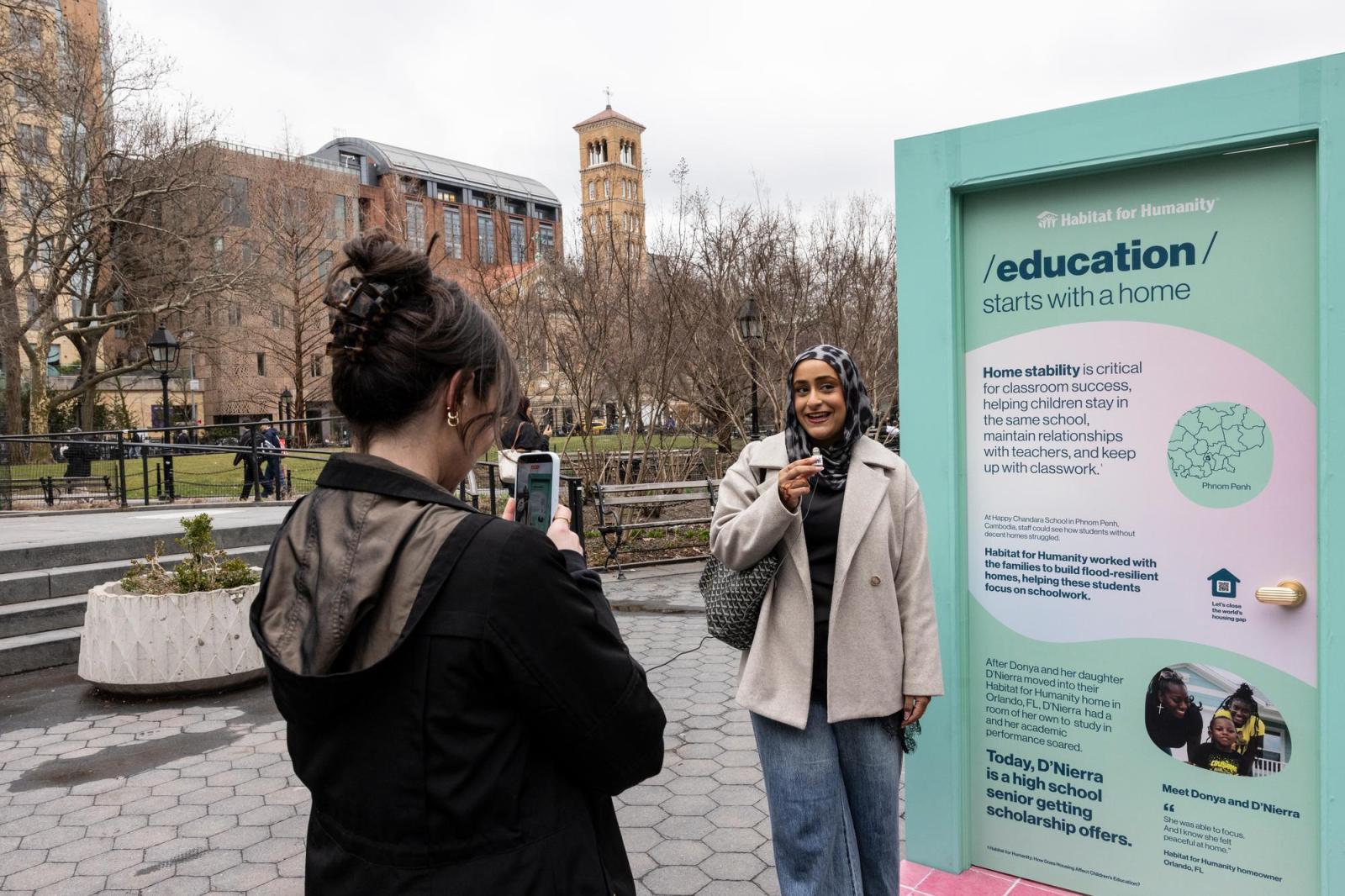 A woman with a hijab and a tan raincoat posing in front of a mint-colored door for her friend to take a photo.