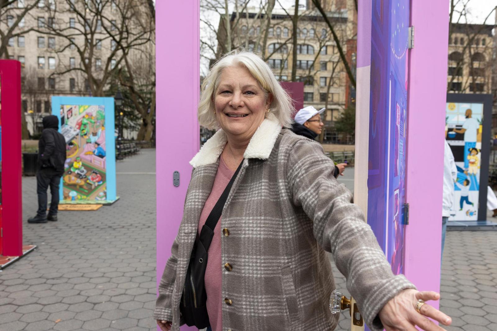 A woman with white hair and a tan plaid jacket walks through a door with a lilac frame.