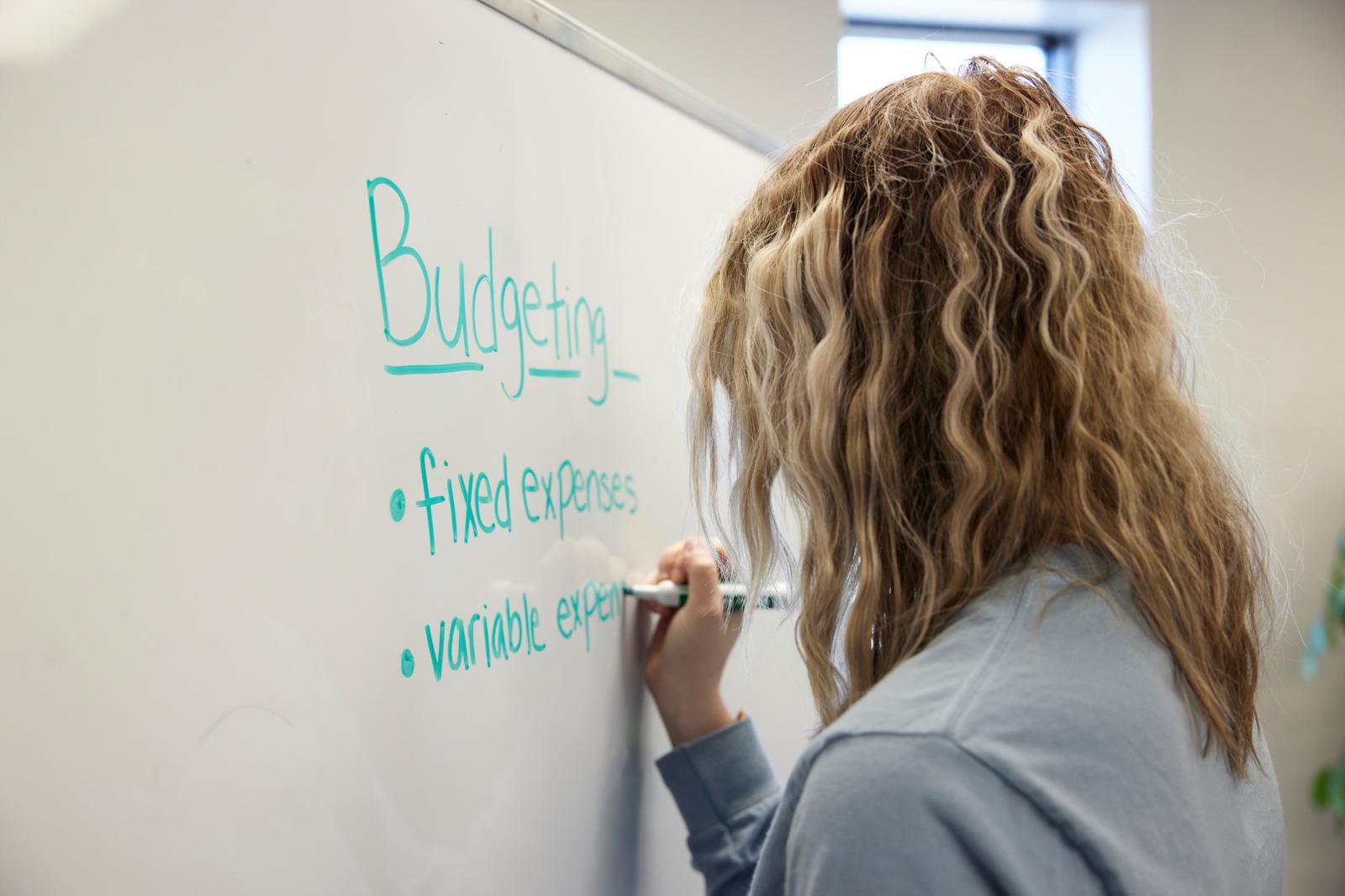 A female instructor with blond hair writing on a whiteboard about budgeting.