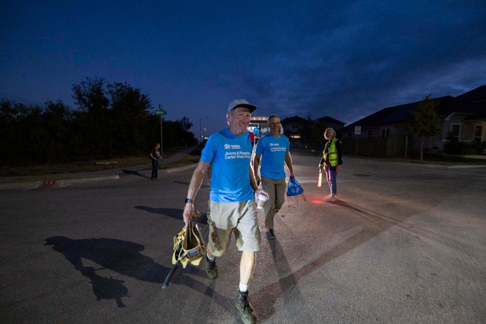 Two volunteers in blue shirts and hats carry tool bags as they walk to the build site before the sun comes up.