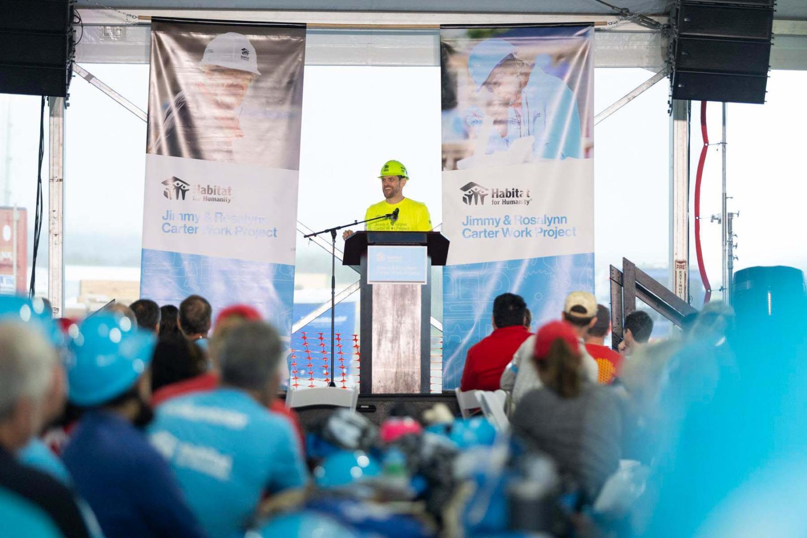John Poynor, home repair project manager, Austin Habitat for Humanity, stands behind a podium on stage in a yellow shirt and white hard hat while the crowd listens. 