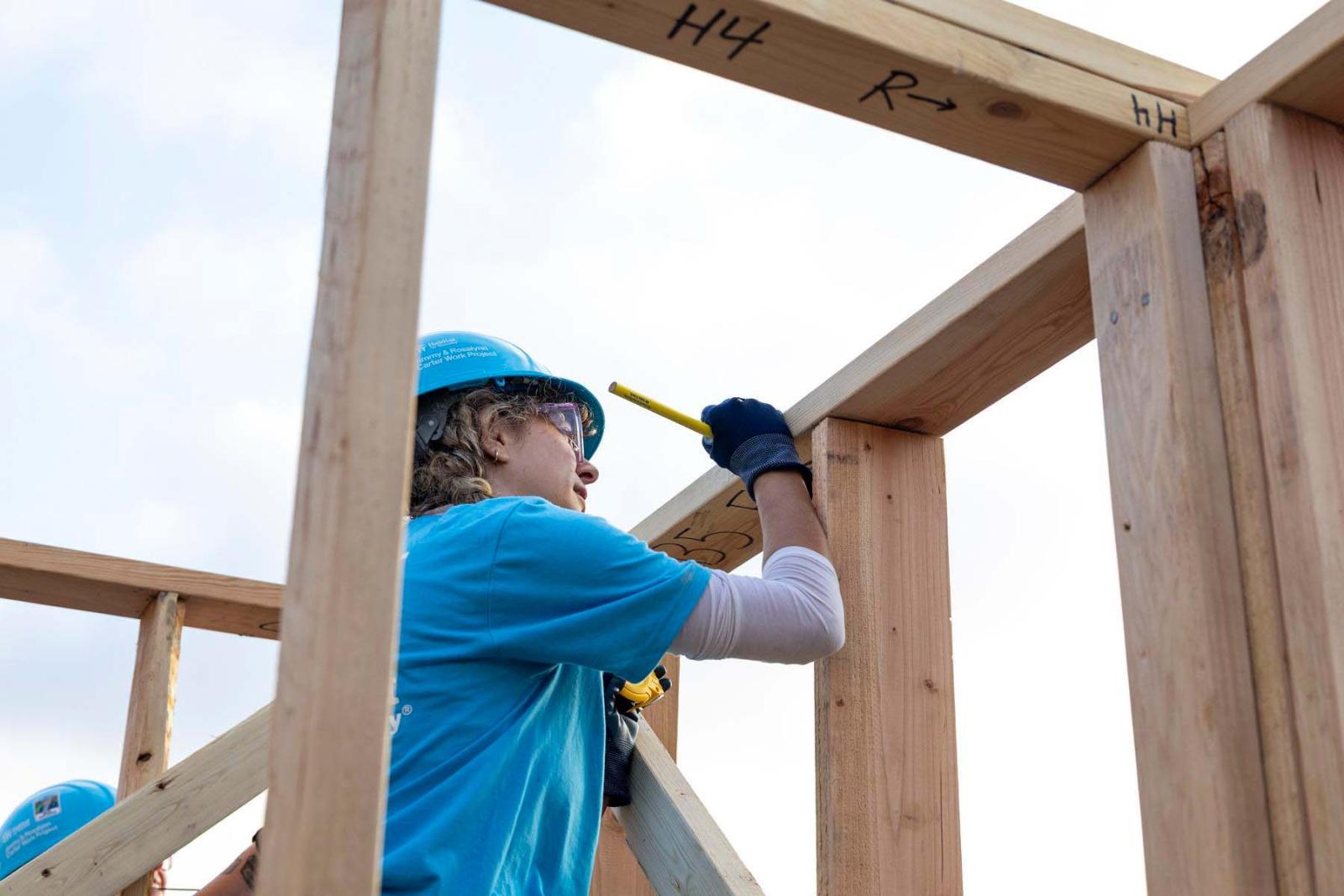 A volunteer in a long sleeve white shirt and blue Habitat shirt and hard hat stands on a ladder marking a piece of lumber with a pencil.