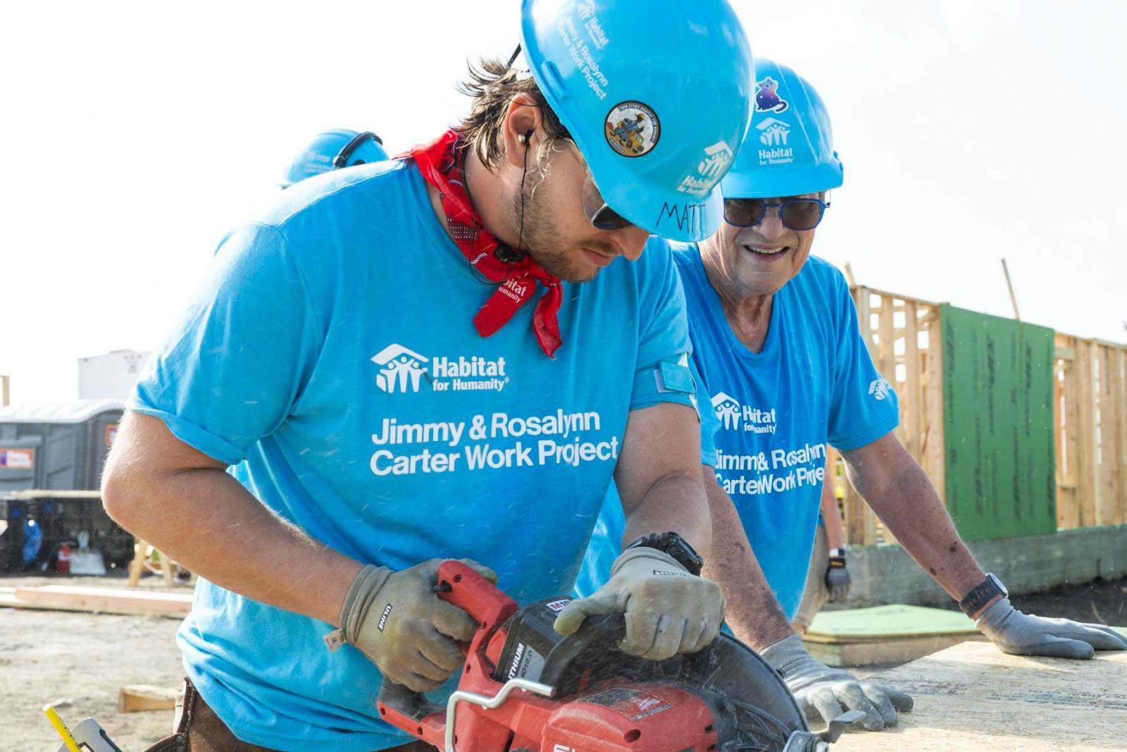 One volunteer in a blue Habitat shirt and hard hat uses a red power saw while the other volunteer observes.