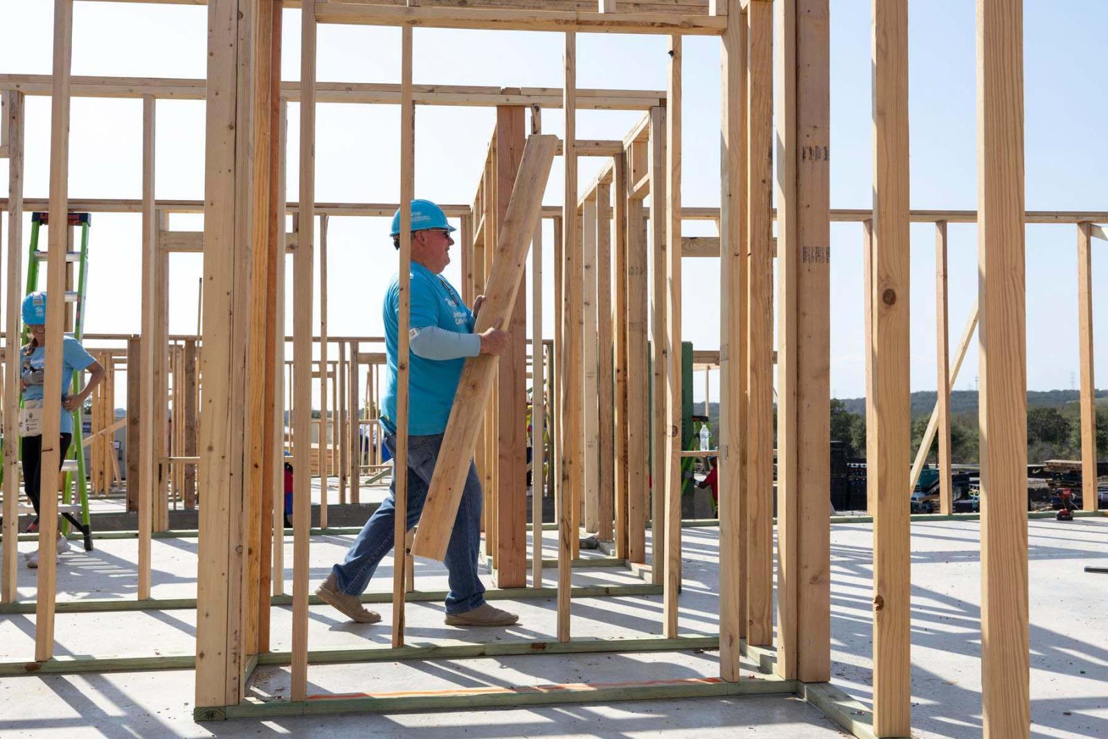A volunteer in a hard hat carries an armload of lumber through the open wooden frame of a house.