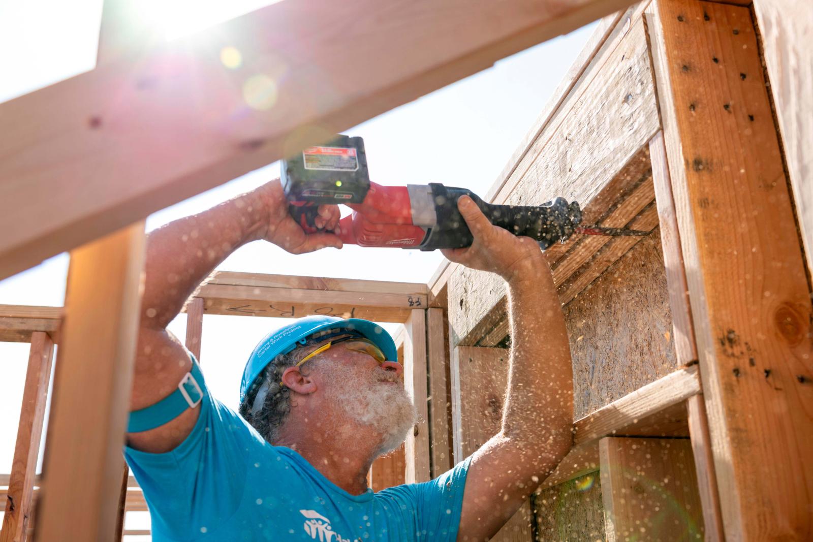 A volunteer stands with his arms above his head holding a drill that he is pushing into the frame of a house.