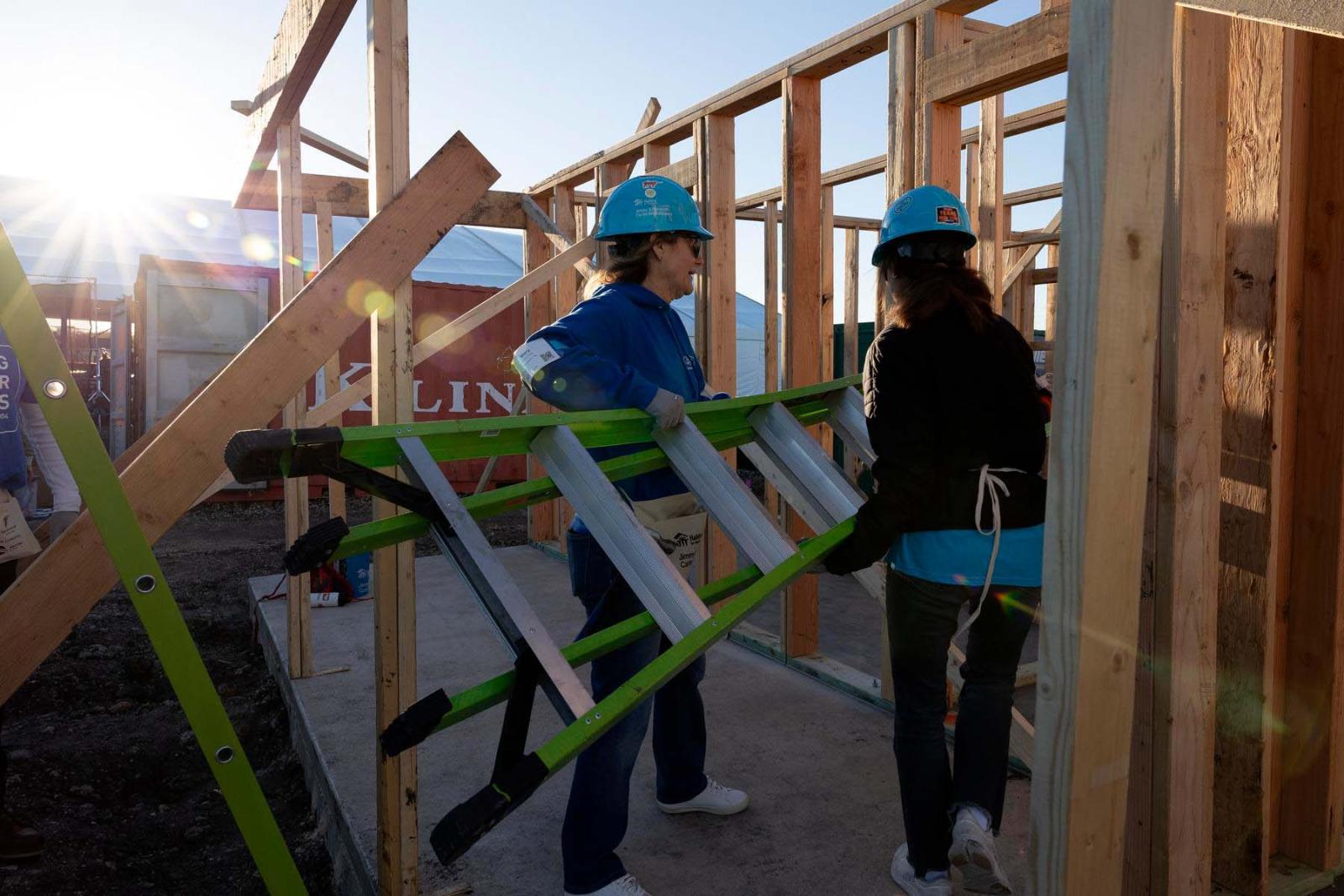 Two volunteers in blue Habitat hard hats hold a green ladder between them as they stand in the wooden frame of a house.