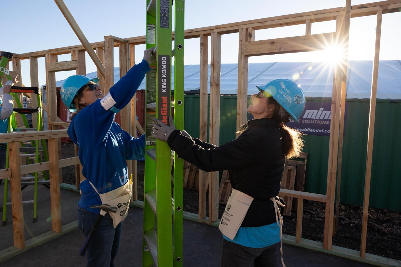 Two volunteers hold up a ladder in the middle of a framed-out house build while wearing hard hats.