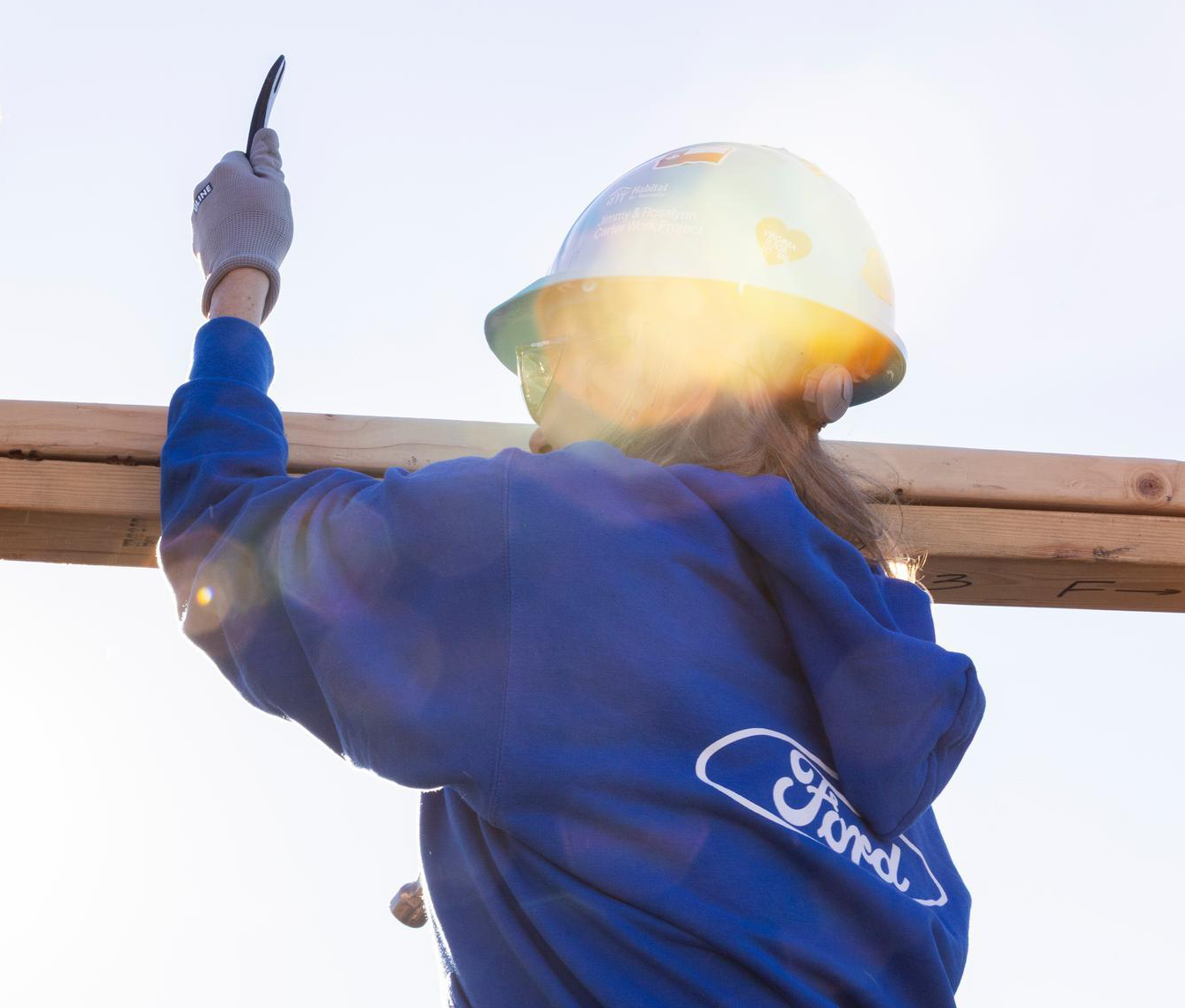 A woman in a blue hoodie with a Ford logo on it and a white hard had stands in a house frame.