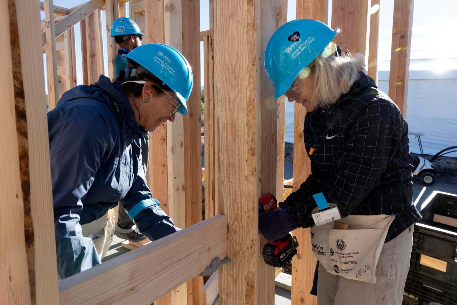 Two volunteers in blue hard hats lean over to drill a piece of lumber that is attached to the wooden frame of a house.