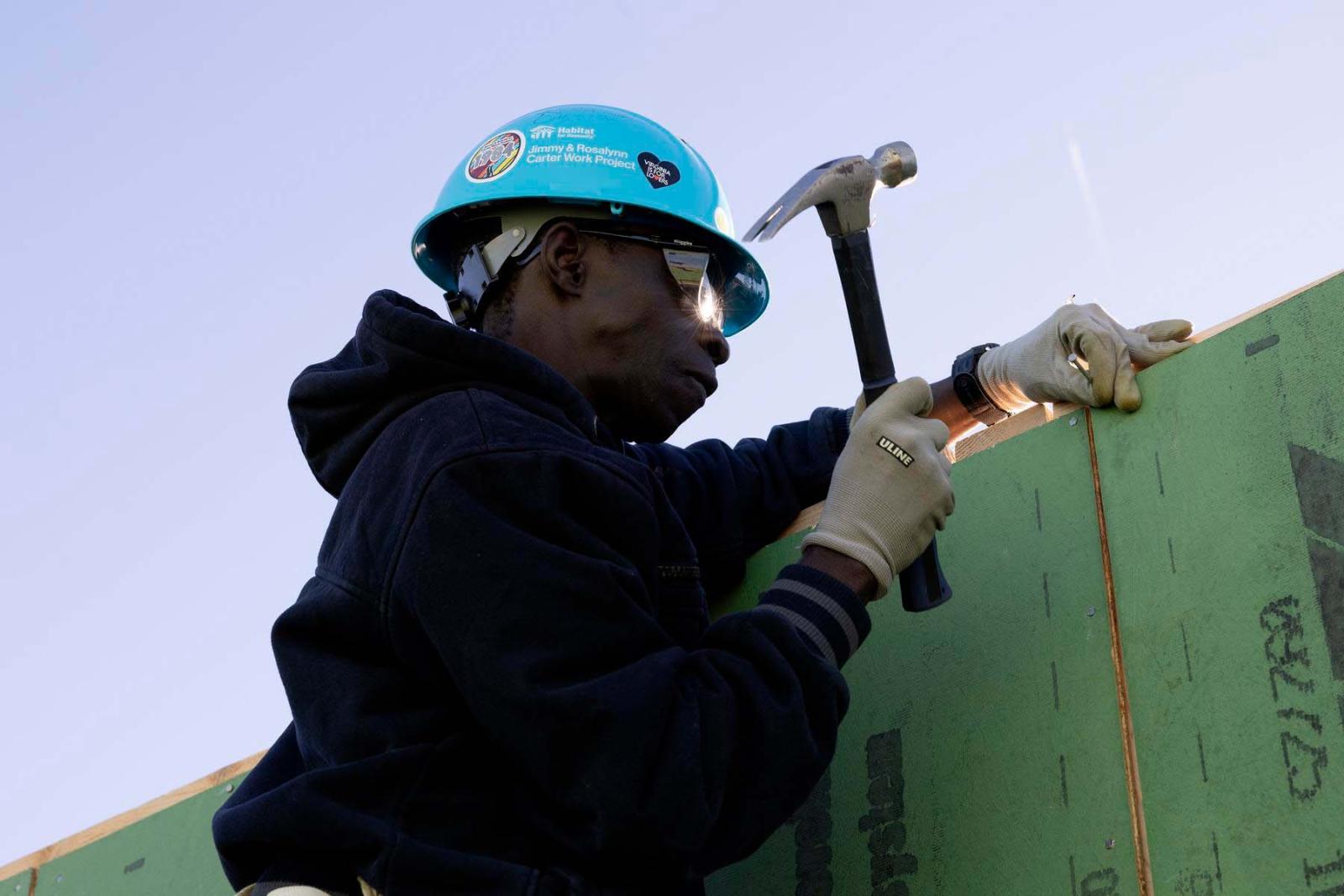 A volunteer in a blue hard hat stands with a green piece of boarding in his hand and a hammer in the other hand.