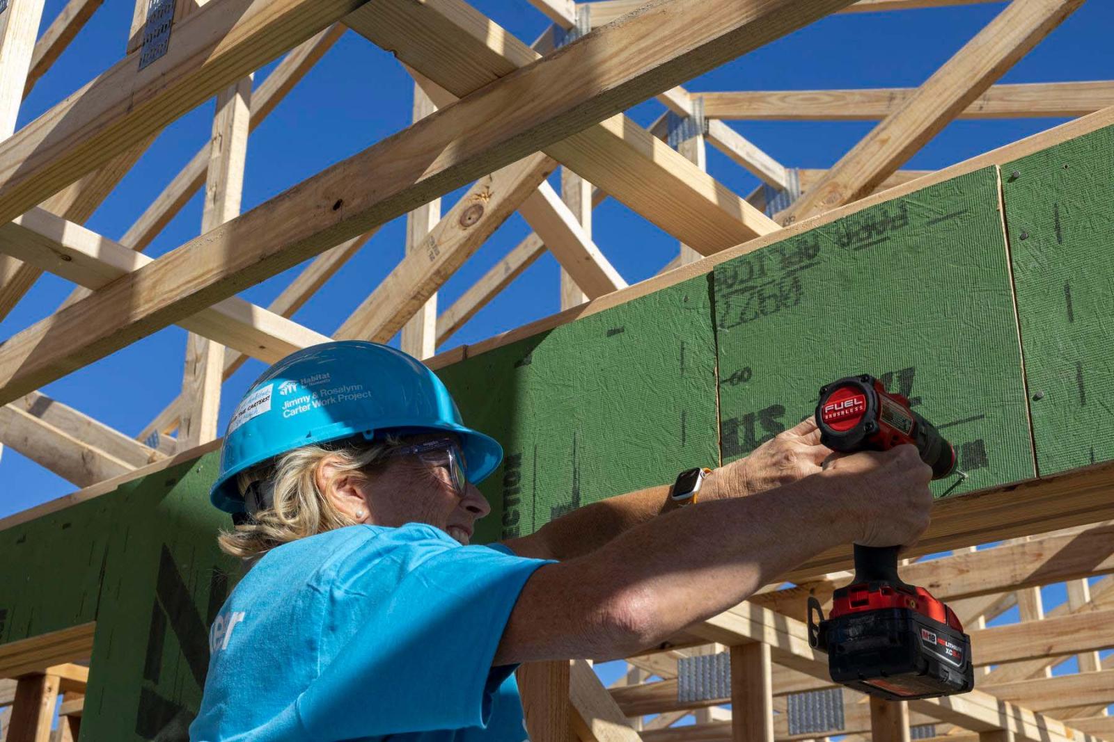 A volunteer in a blue hard hat and shirt works with a drill in front of green drywall.