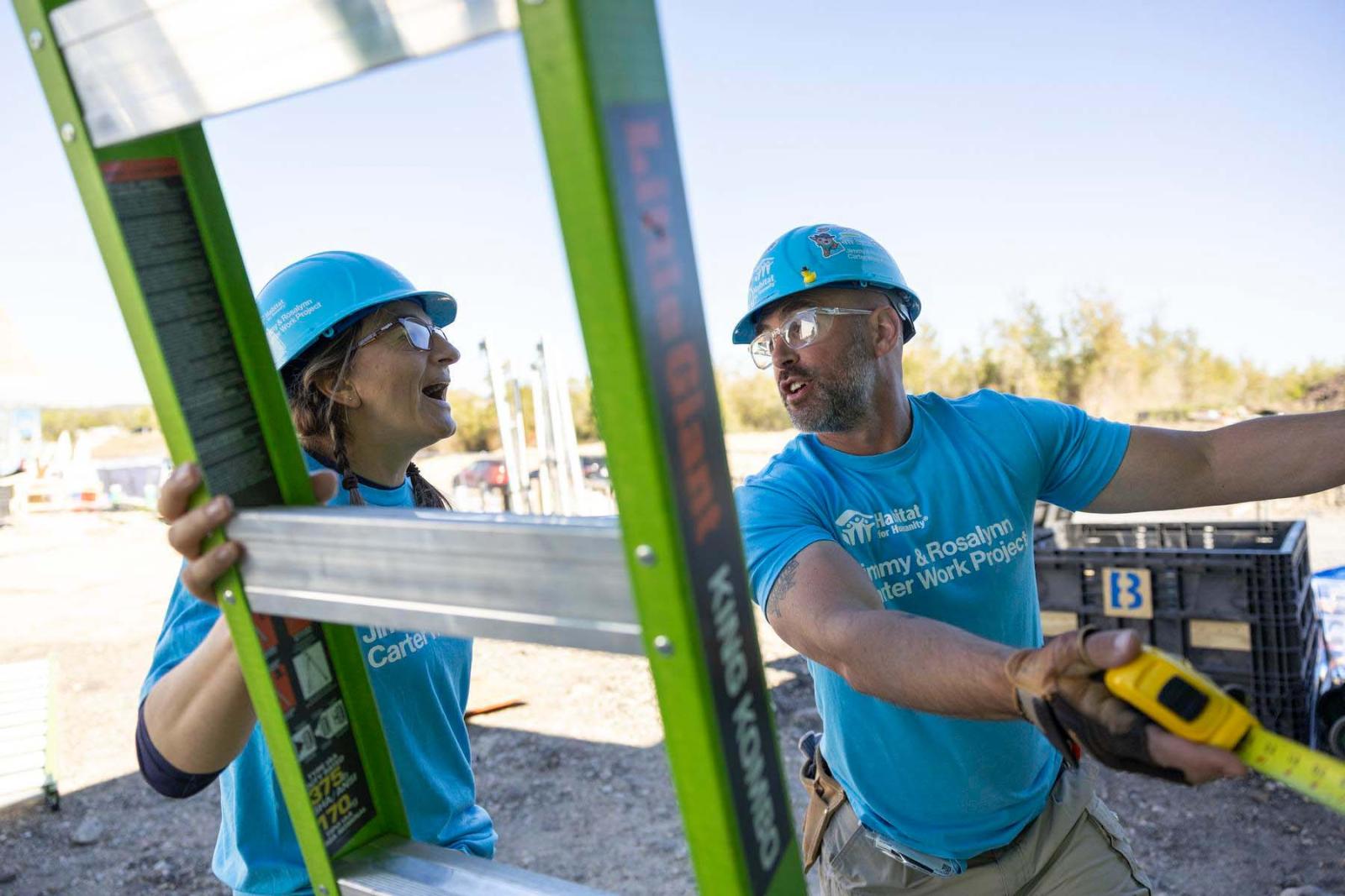 Two volunteers in blue shirts and hard hats stand with a green ladder between them and holding a measuring tape.