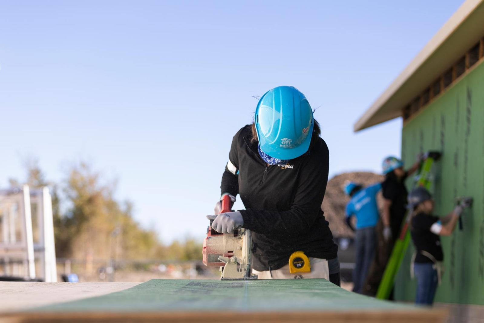 A volunteer in a blue hard hat leans forward and uses a handsaw on a piece of lumber.