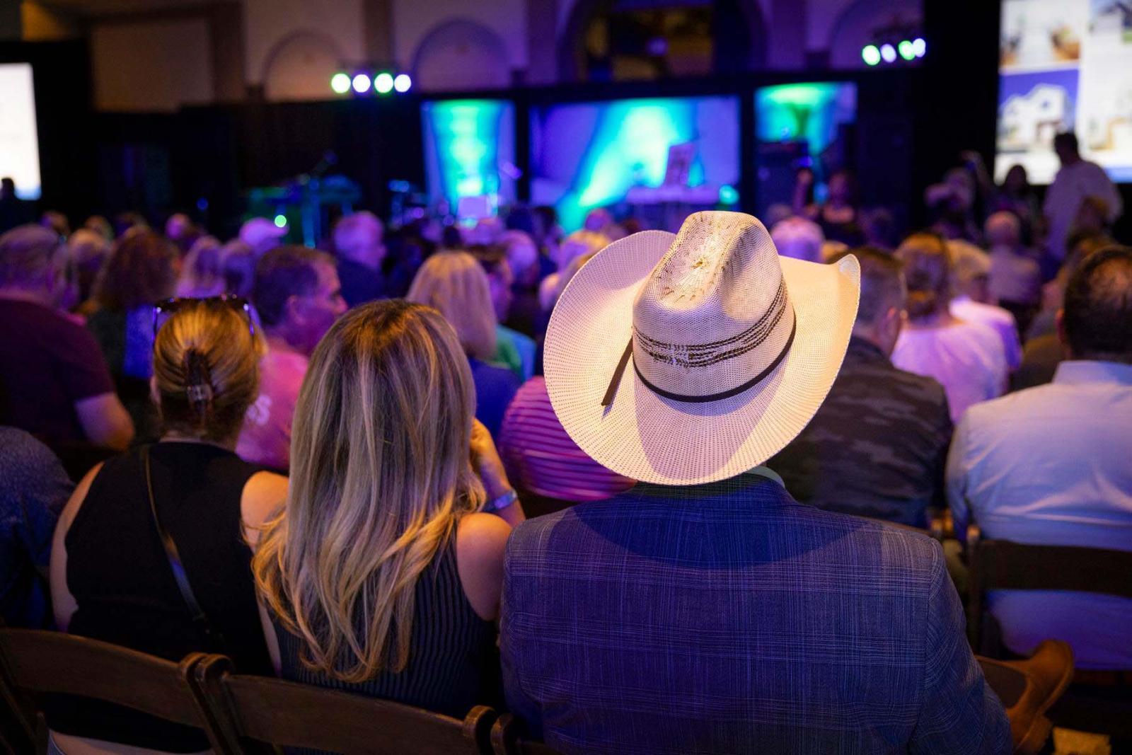 The back of the audience's heads, including someone wearing a large white cowboy hat, are displayed as they watch the stage for the opening ceremony. 