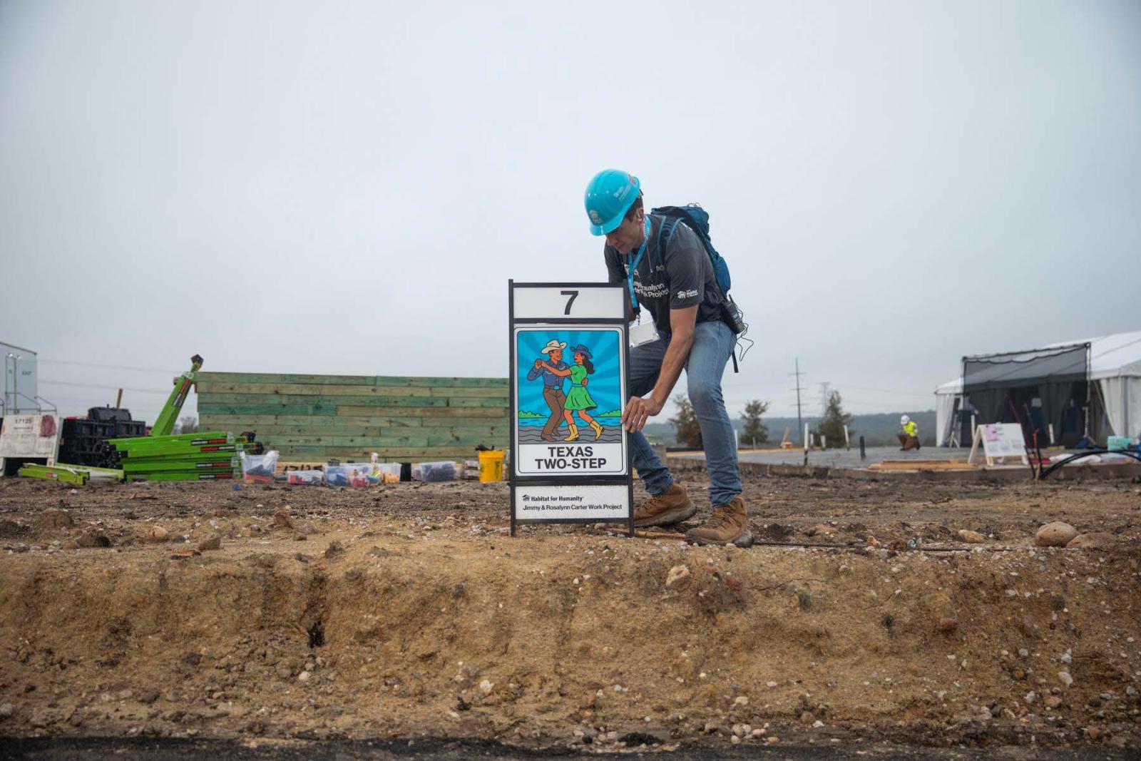 A man in a blue hard hat sets up a sign that has a dancing man and woman on it that says Texas Two-Step.