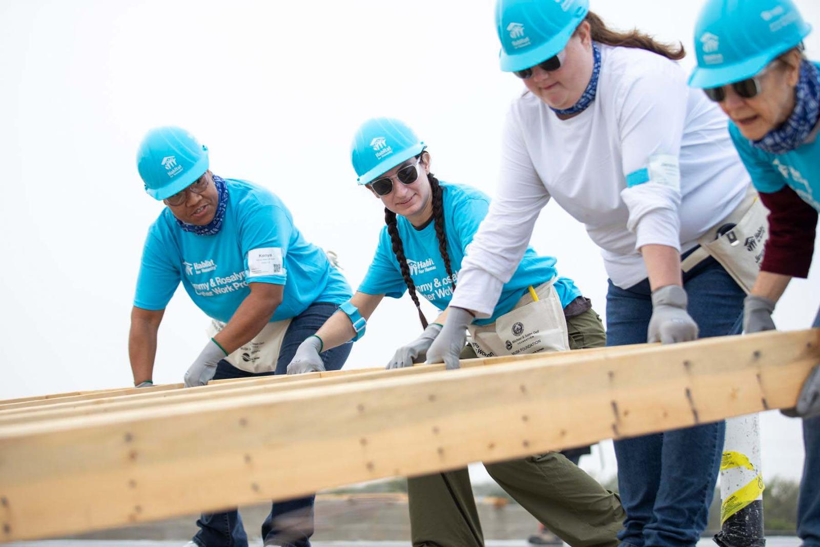 Four volunteers in blue hard hats pull on a wooden frame wall of a house to lift it up off the ground.