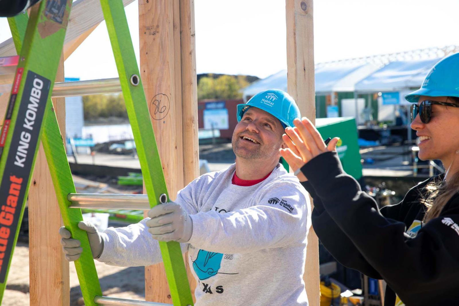 A volunteer in a white shirt and blue hard hat holds the middle of a green ladder steady and looks upward.