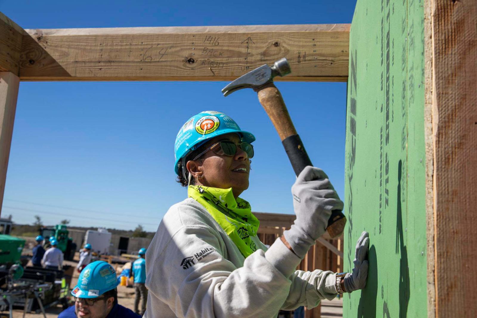 A volunteer with a blue hard hat holds a hammer in their hand getting ready to hammer a nail in to place on a wall.