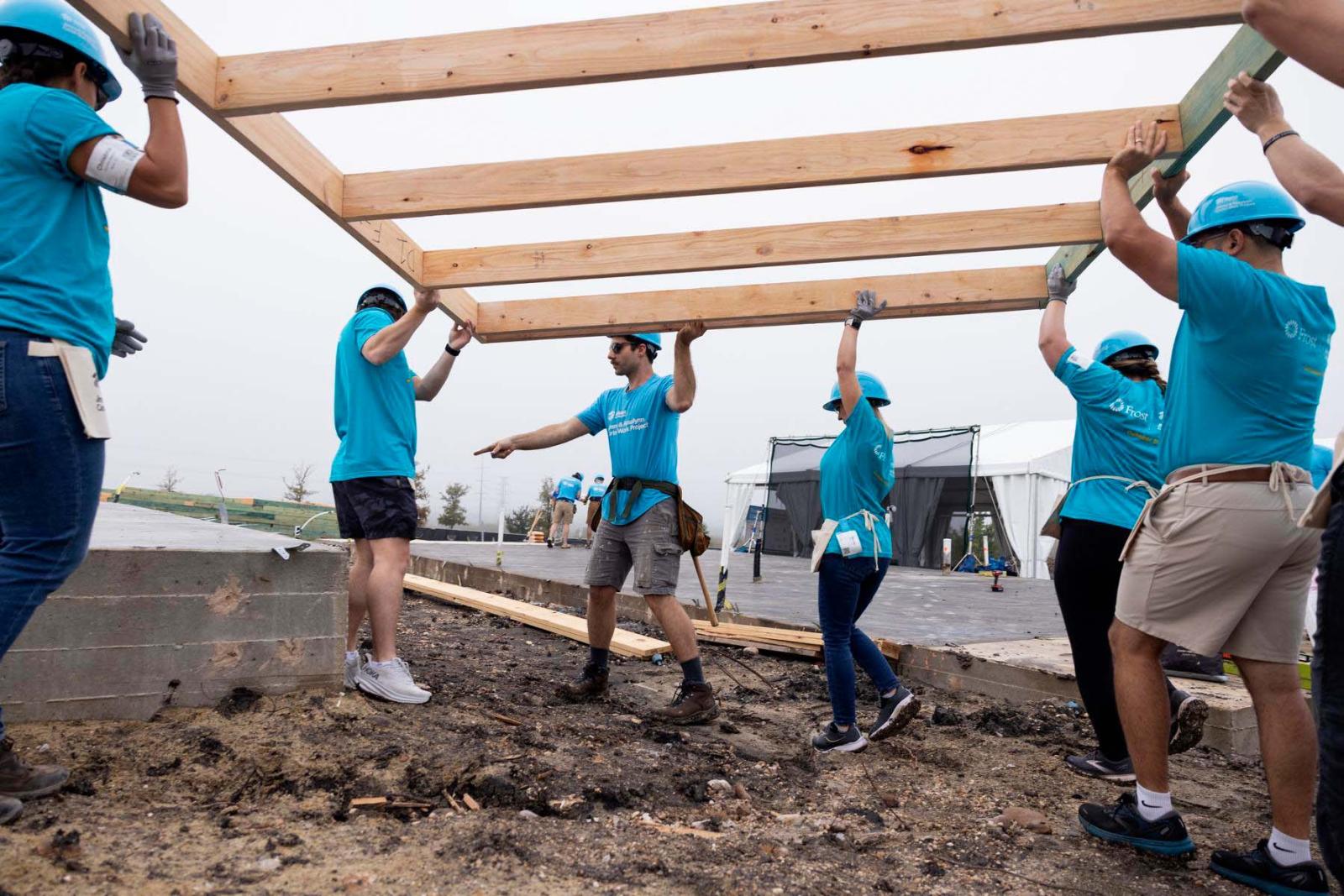 Seven volunteers wearing blue Habitat shirts lift a wooden frame above their heads