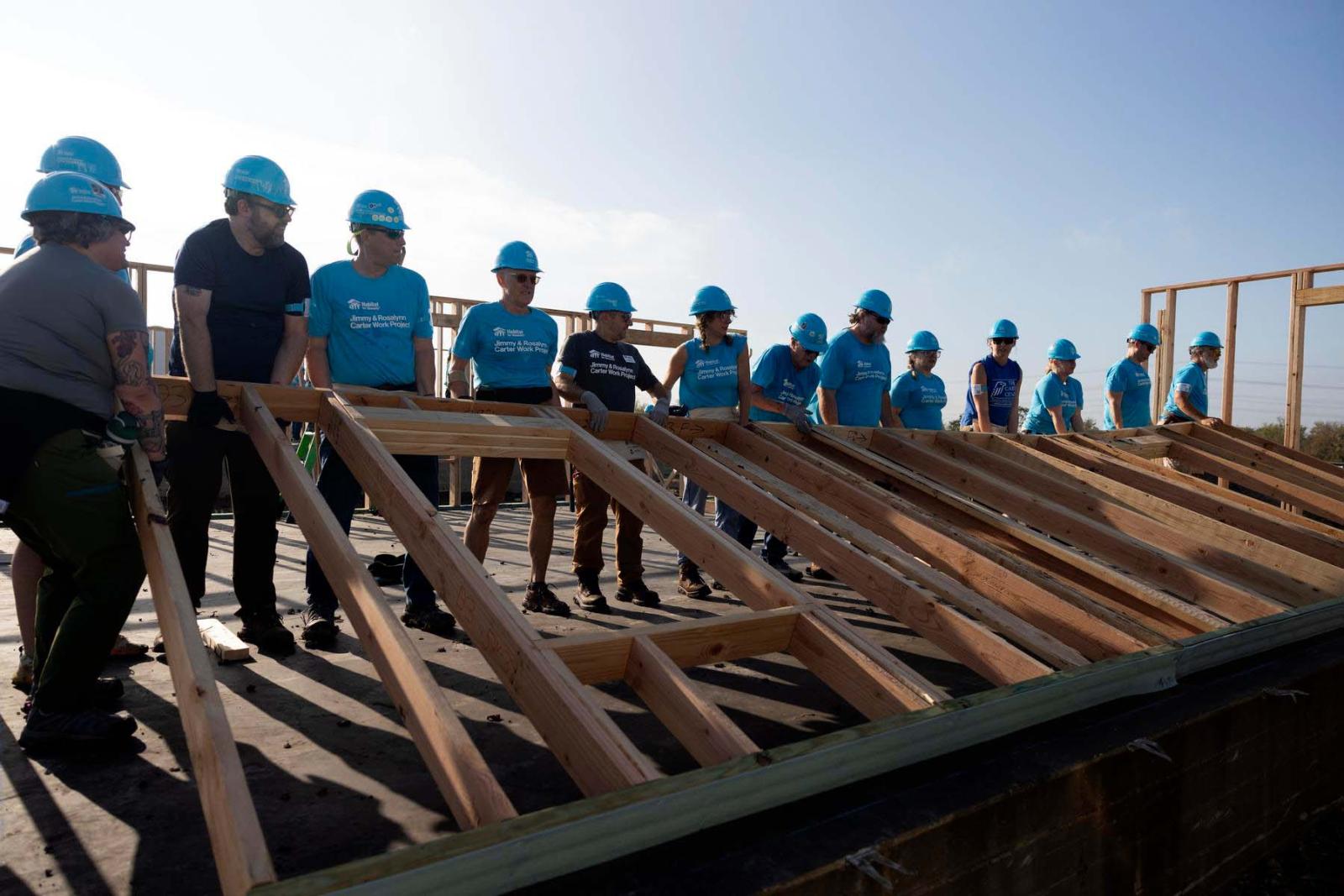 Thirteen volunteers in blue Habitat shirts and hard hats stand on one side of a wooden frame of a house preparing to lift it into place.