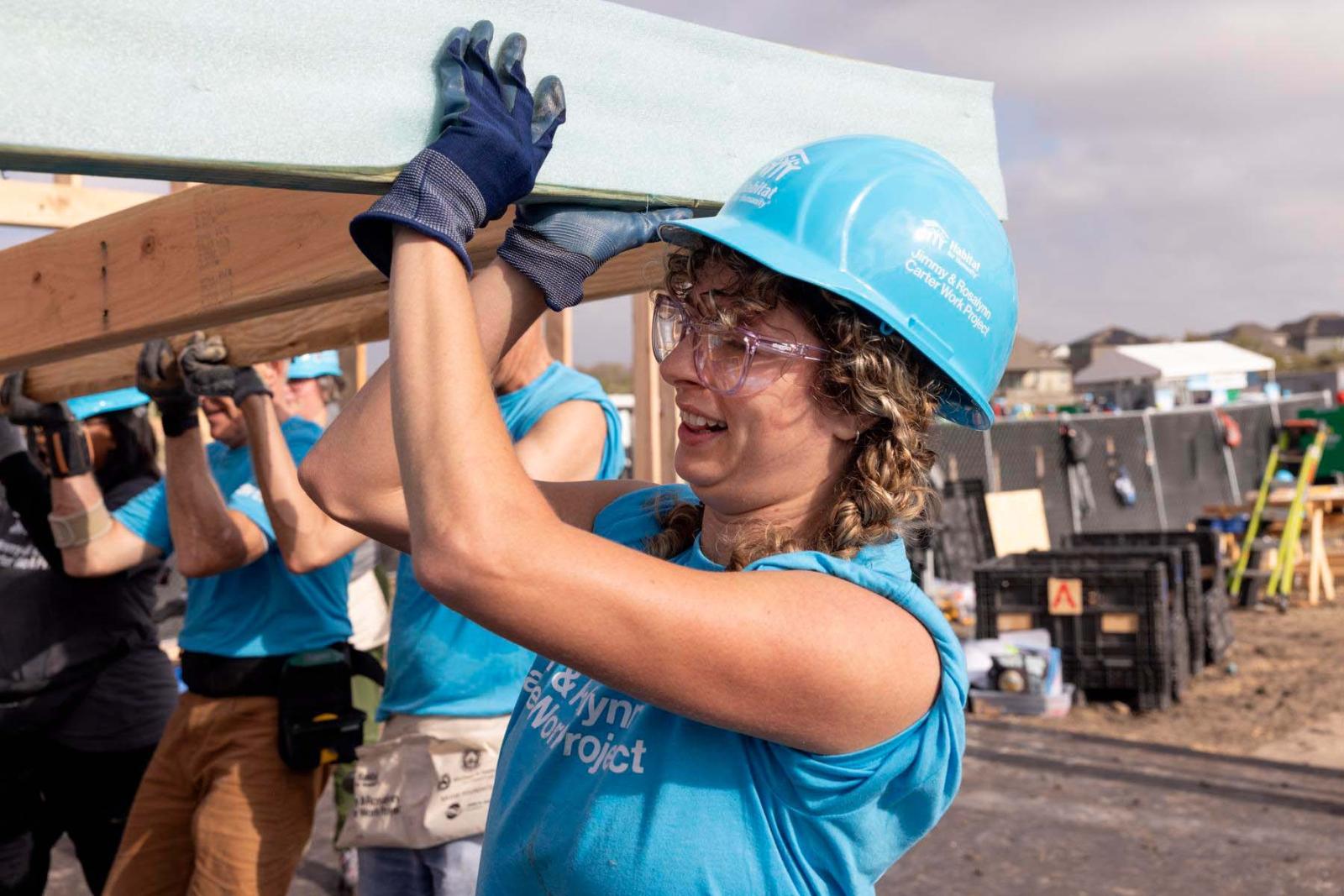 A volunteer with curly hair wearing a blue Habitat shirt and hard hat lifts a piece of lumber above her head.