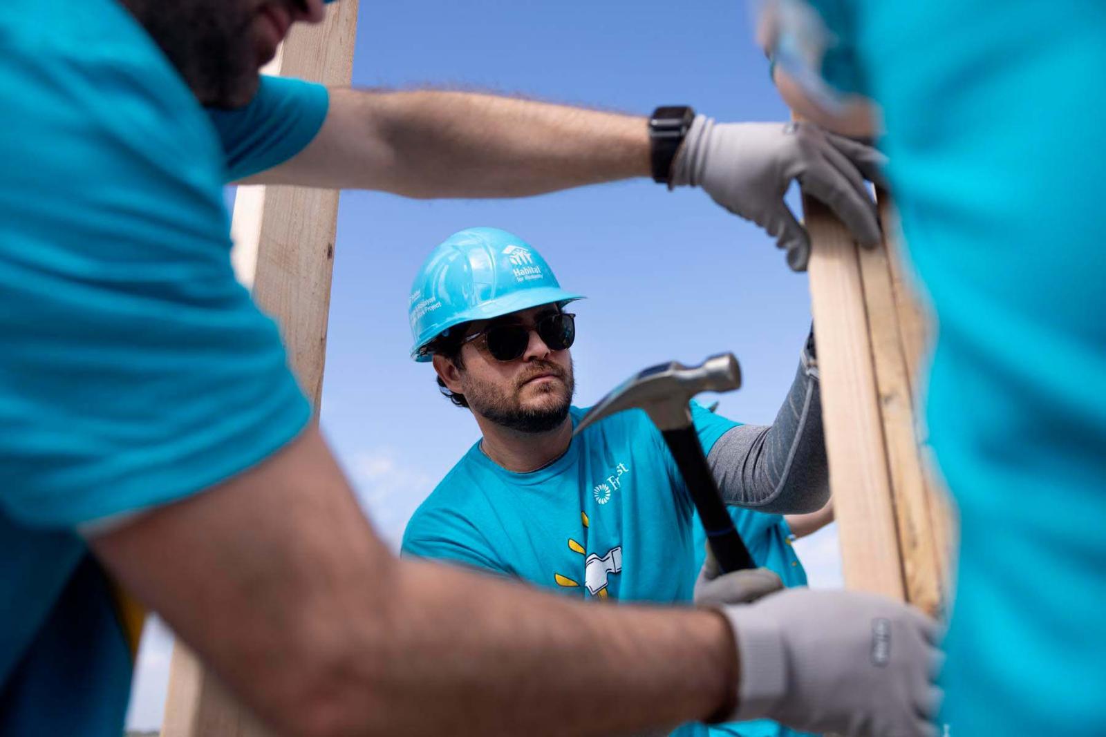 In the background a volunteer in a blue hard hat hammers a piece of lumber. His face is framed by another volunteer's arms working in the foreground.