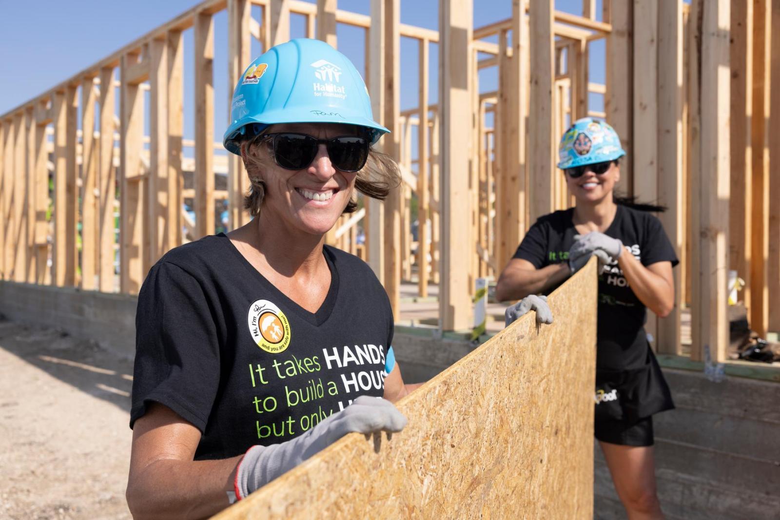 A volunteer in a black t-shirt and blue hard hat holds a piece of lumber and smiles directly at the camera.