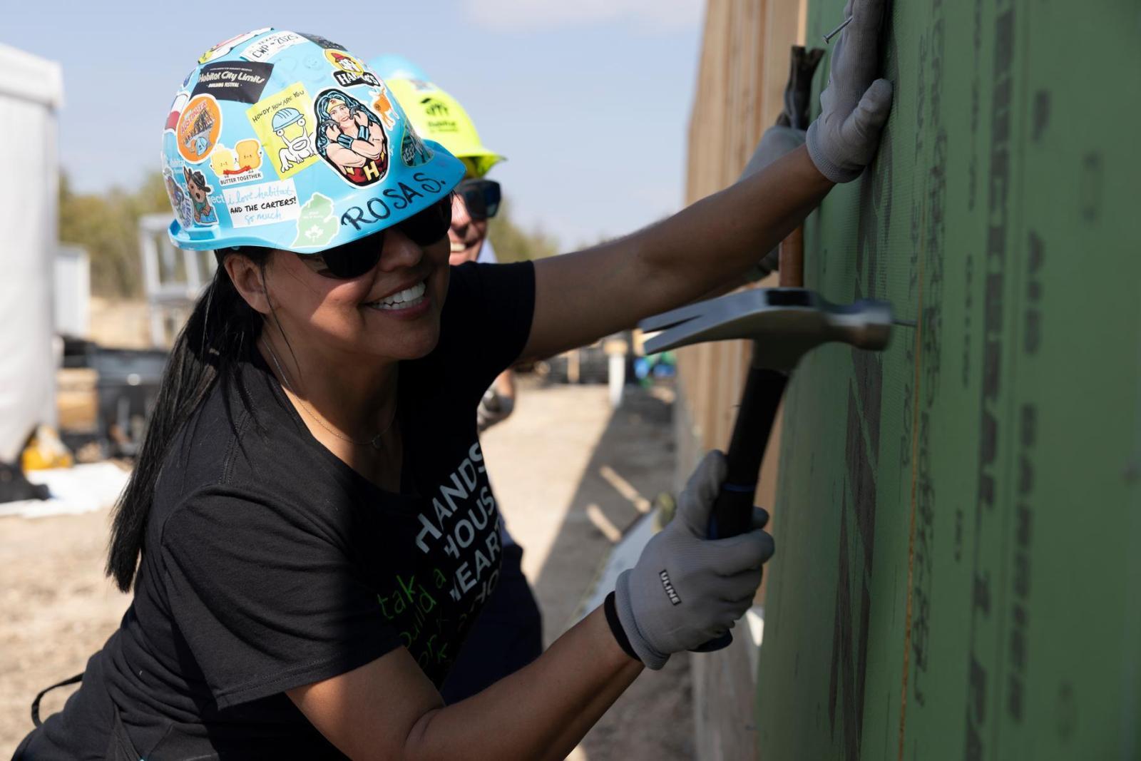A volunteer in a black t-shirt and blue hard hat holds a hammer and leans toward the wall to get to work.