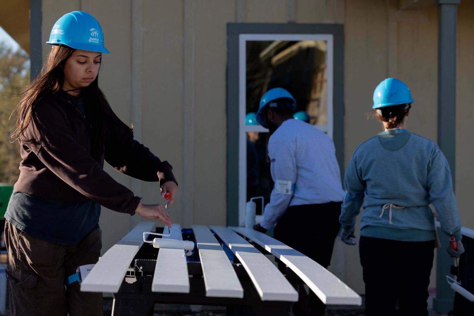 Three volunteers in hard hats work on using paint rollers to paint pieces of lumber white.