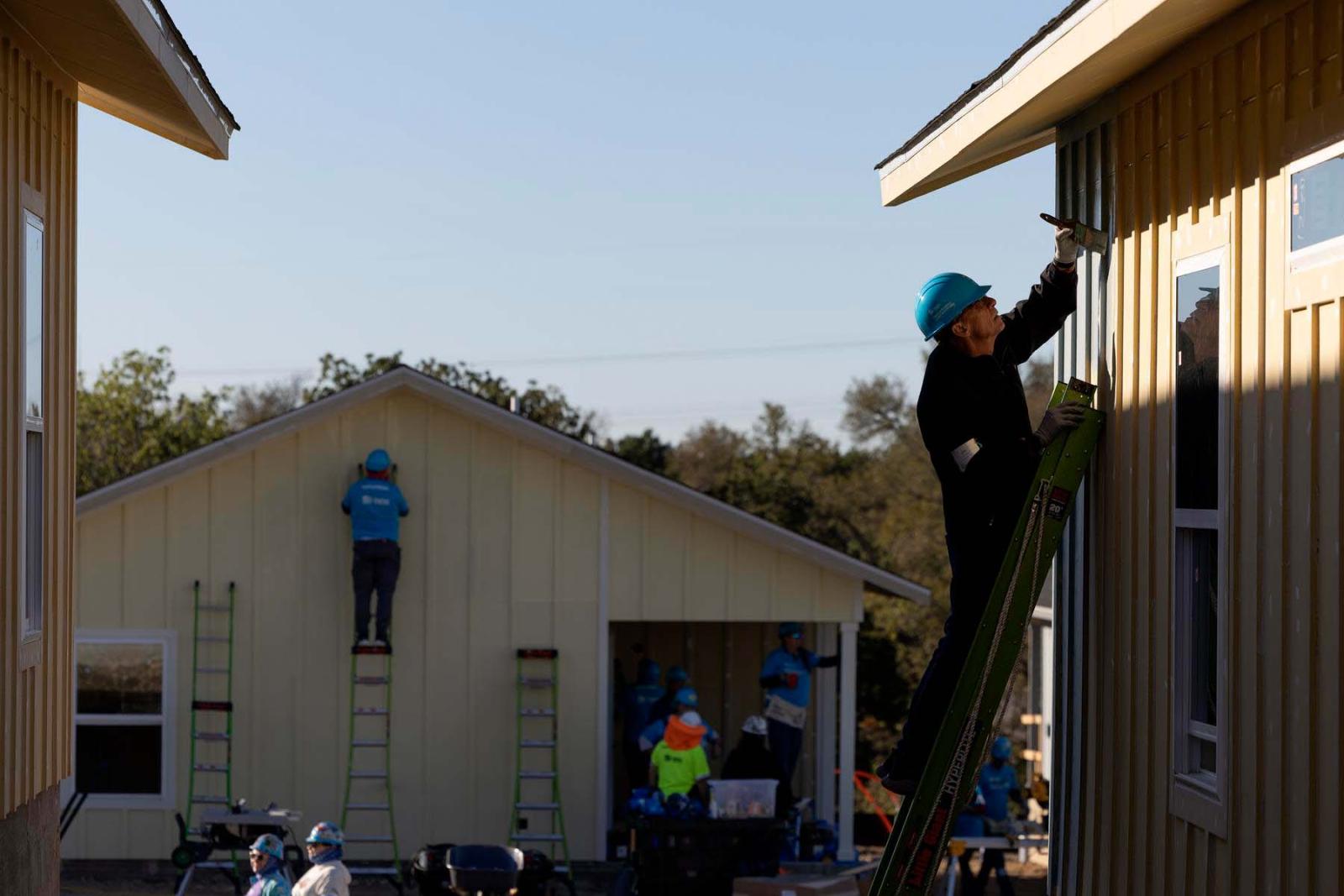 Two volunteers stand at the top of ladders leaning against the outside wall of two separate houses.