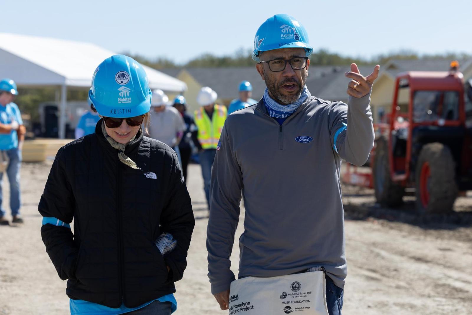 Two people stand in blue Habitat hard hats on a build site discussing the work.