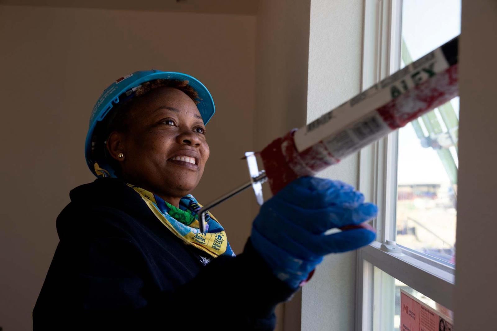 A volunteer in a blue Habitat hard hat uses a caulking gun on a window frame inside a Habitat house.