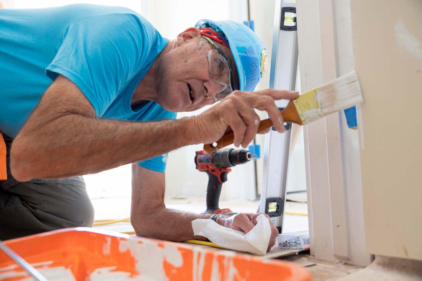 A volunteer in a blue hard hat bends down and angles his arm in front of him holding a paint brush as he paints a wall white.