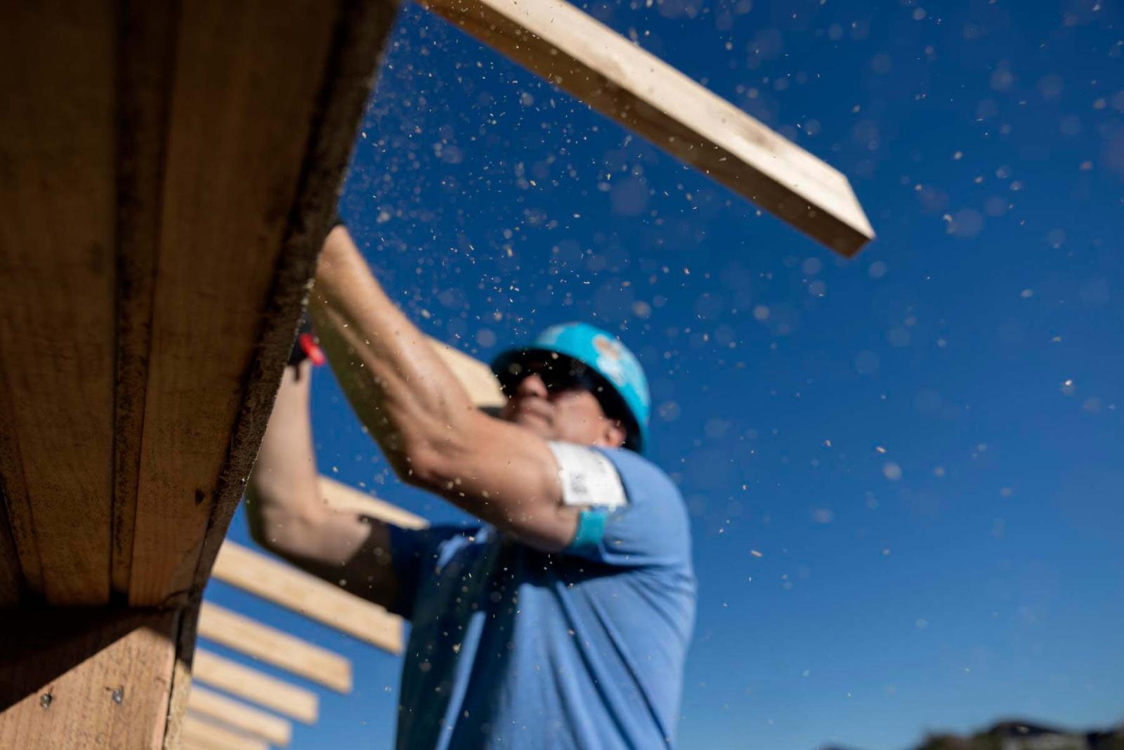 A volunteer in a blue shirt and hard hat stands on a ladder and reaches his arms out to the top a roof as sawdust falls.