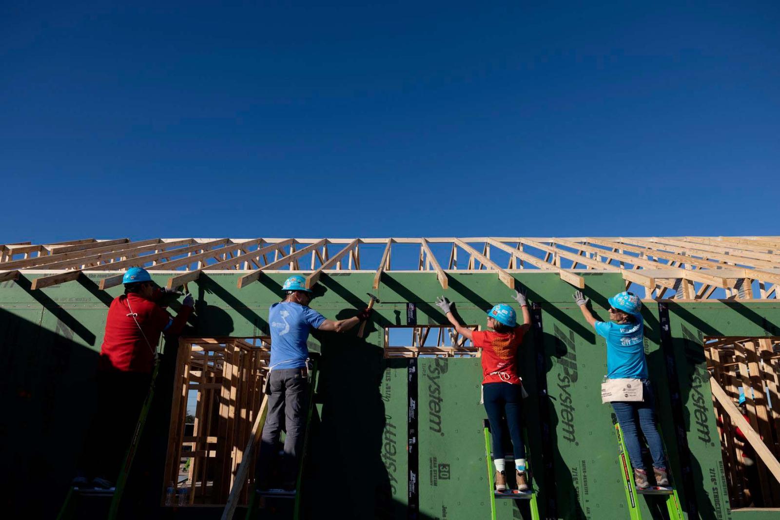Four volunteers in hard hats with their backs to the camera stand on ladders while working on a house frame.